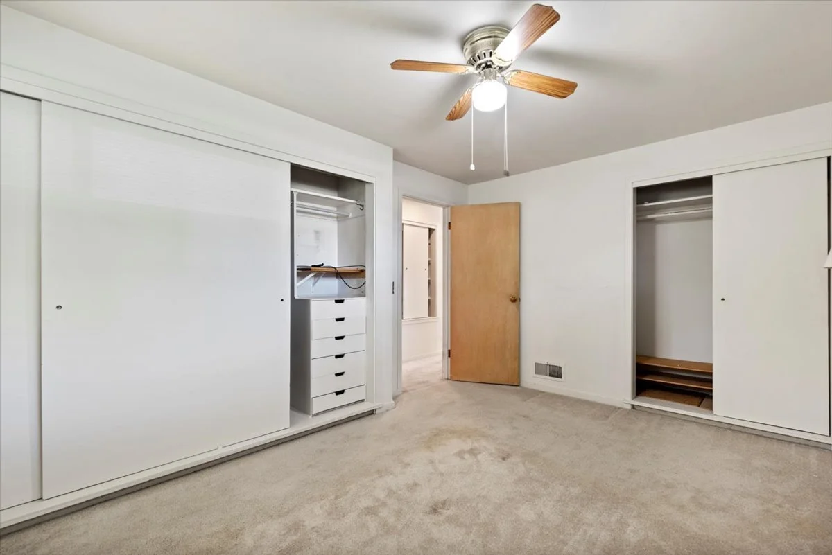 Empty bedroom with beige carpet, white walls, ceiling fan with wooden blades, open closet doors, and a wooden door leading to another room.