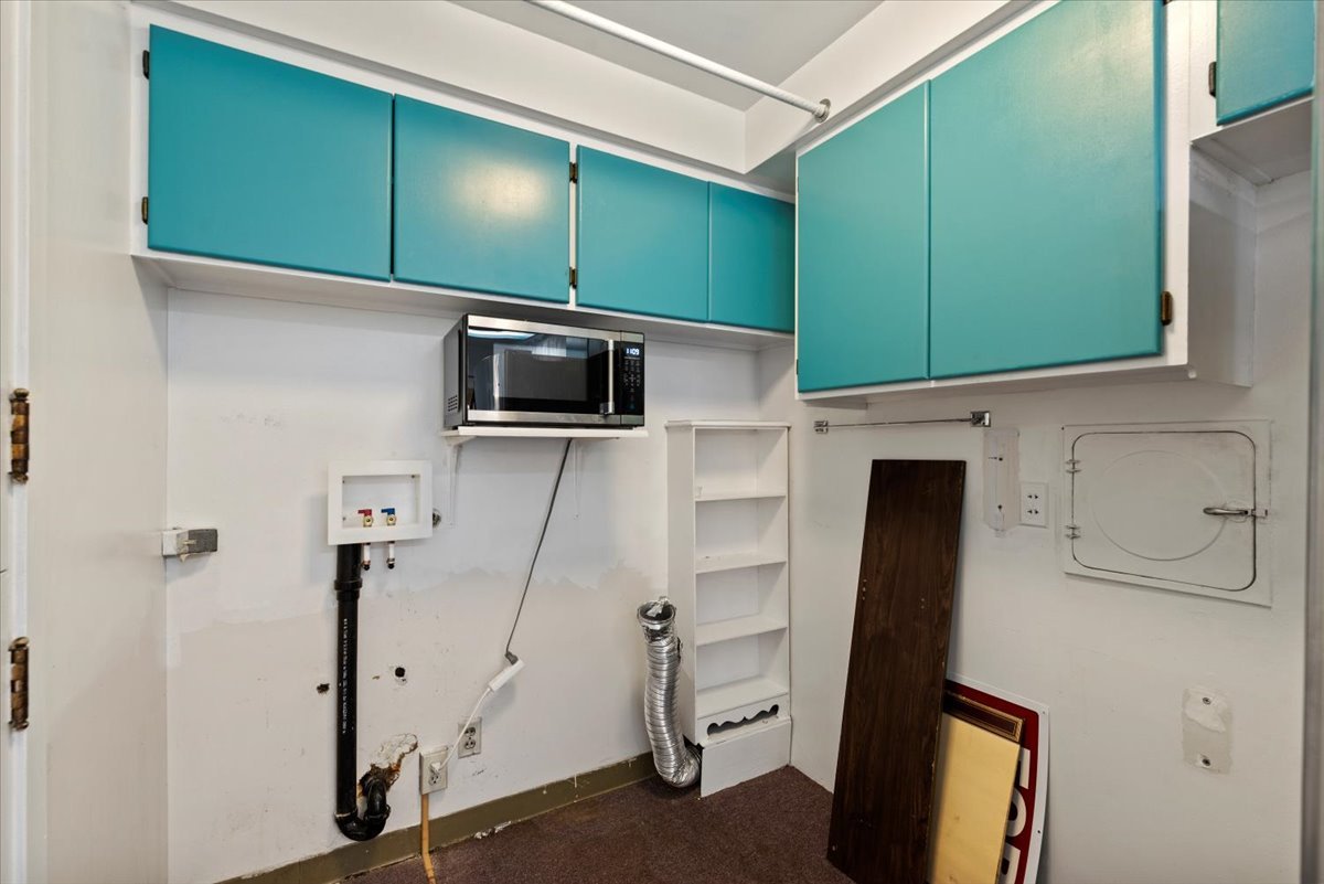 An empty laundry room with teal cabinets on the wall, a microwave on a shelf, and a small white shelf unit. There is a metal duct, an electrical outlet, and a small white access panel on the wall. A wooden board leans against the wall, and a red and 