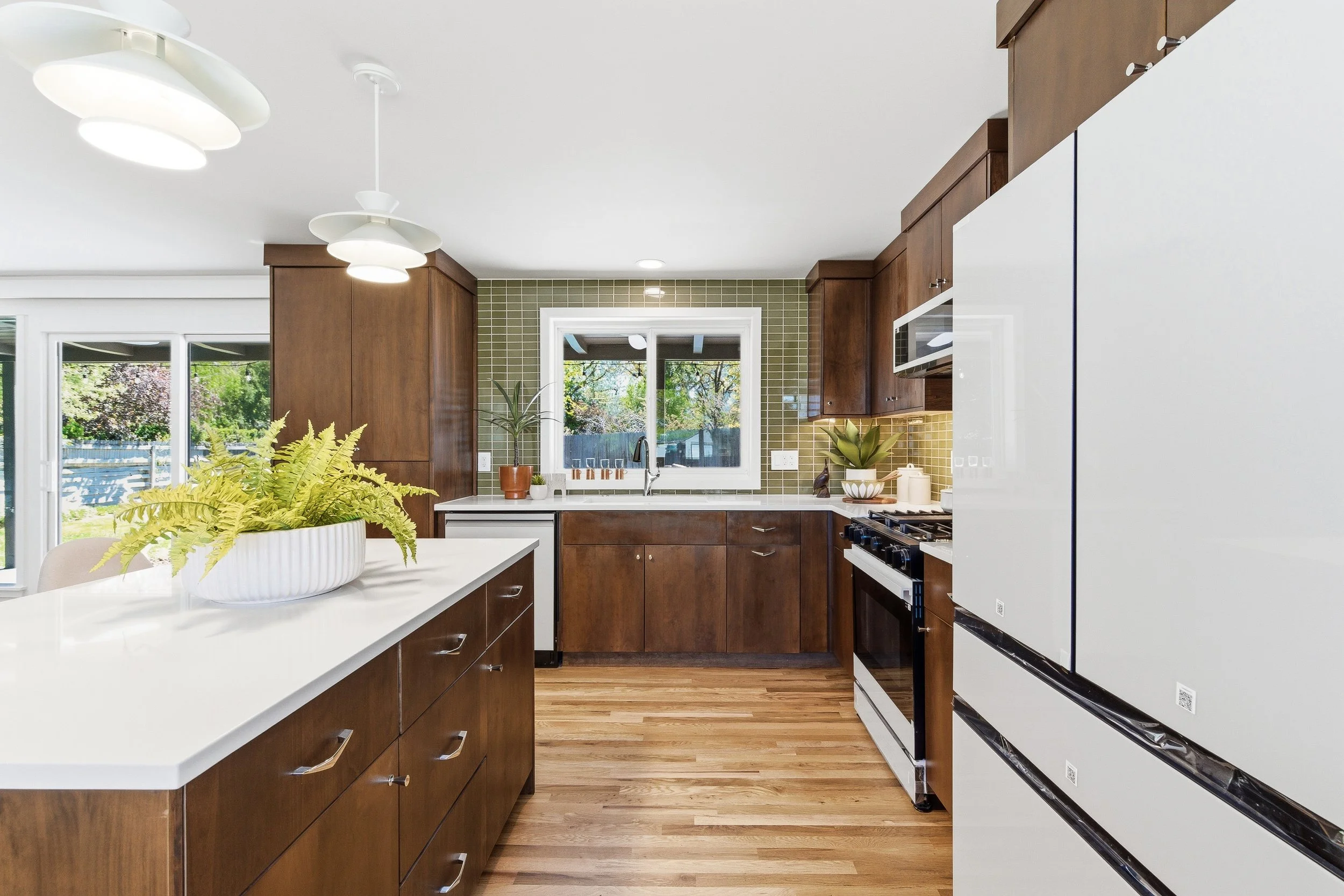 A modern kitchen with wooden cabinets, white countertops, and a window overlooking a backyard. The kitchen features a dining table with a white bowl and green plants, pendant lights, and stainless steel appliances.