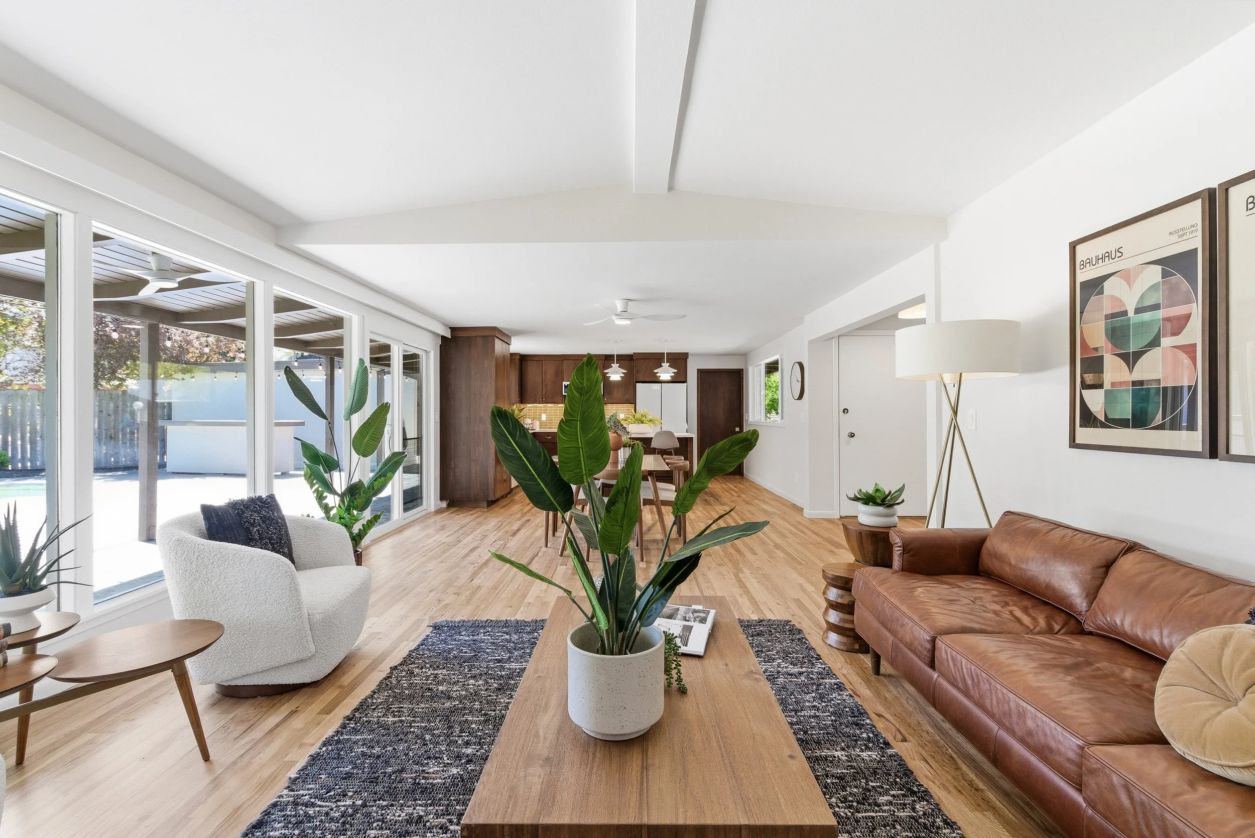A bright living room with wooden flooring, large windows, and sliding glass doors leading outside. There are various green plants, a white armchair, a brown leather sofa, and framed artwork on the white walls. A yellow kitchen with wooden cabinets is