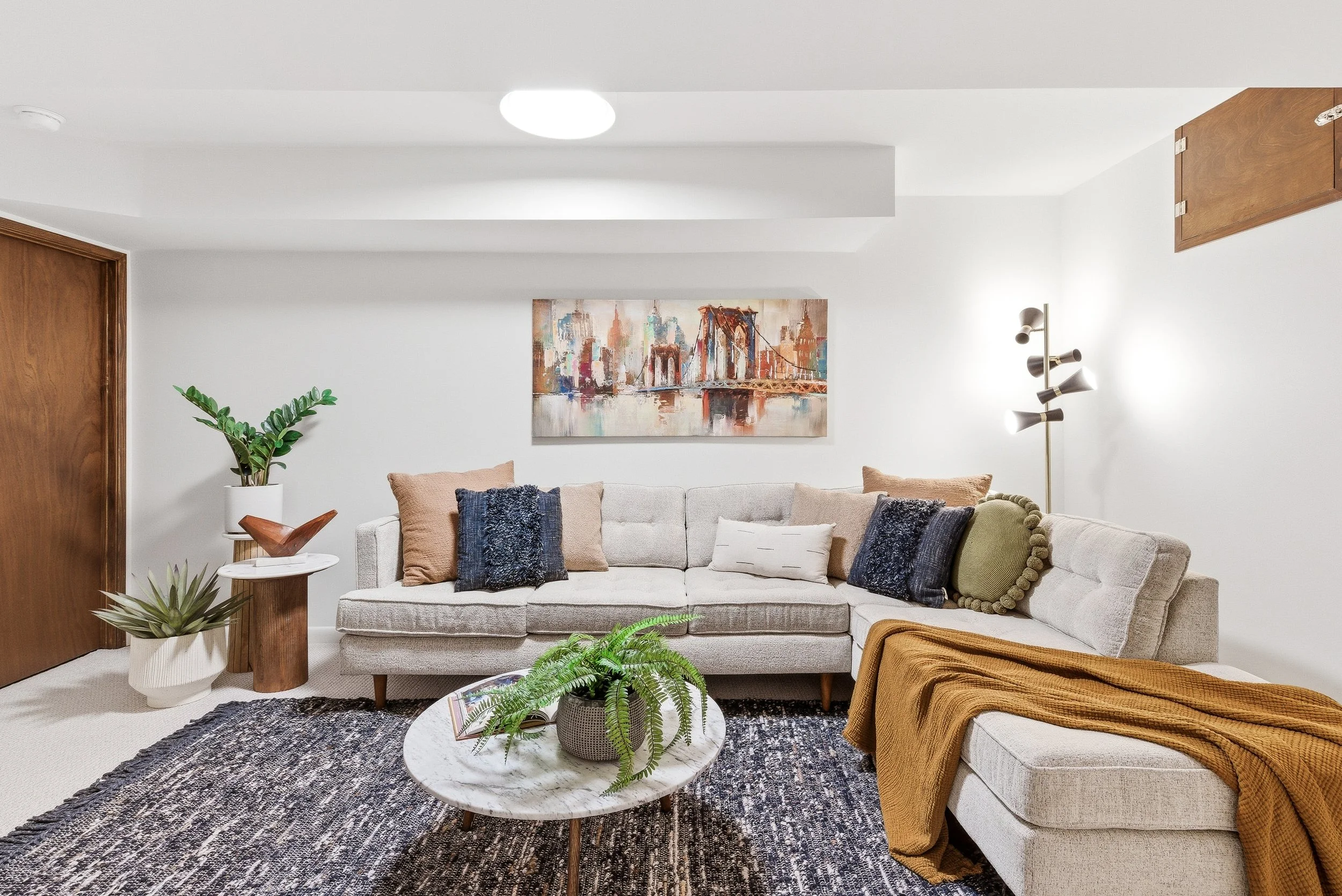 Living room with a beige sofa adorned with various pillows, a white and black patterned rug, wooden side tables, potted plants, abstract cityscape art on the wall, a modern floor lamp, and a wooden ceiling cabinet.