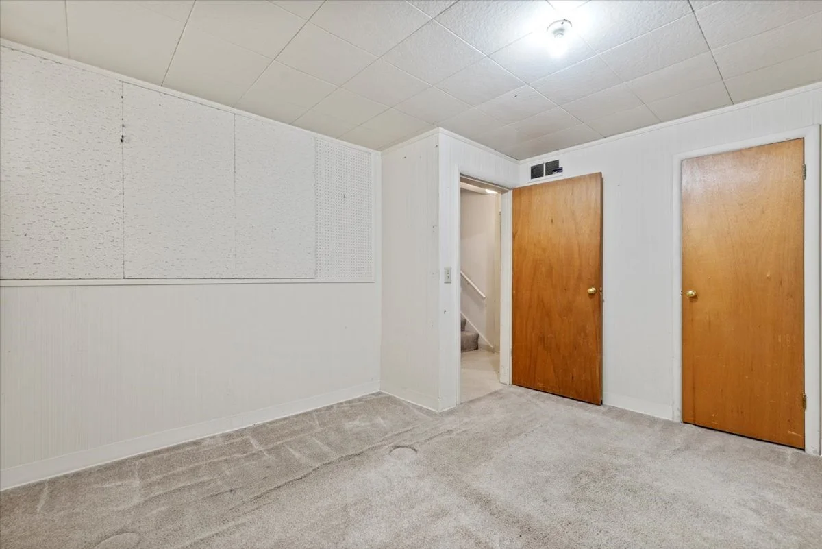 Empty room with beige carpet, white walls, and two wooden doors. One door is slightly open revealing stairs, and the other door is closed. A ceiling light illuminates the room.