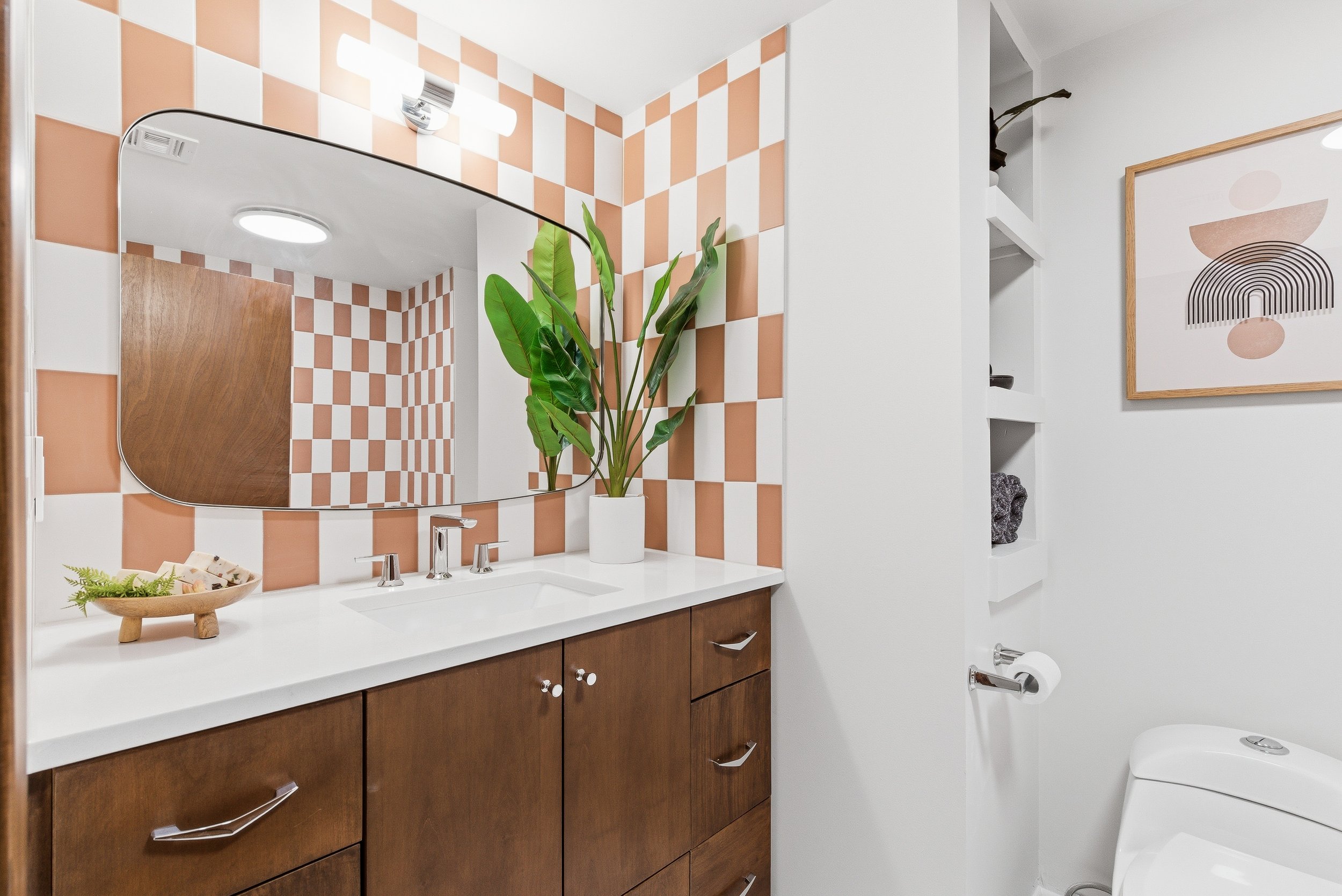 A modern bathroom with a white countertop, wooden cabinets, a large mirror, a potted green plant, and orange and white checkered tiled walls.