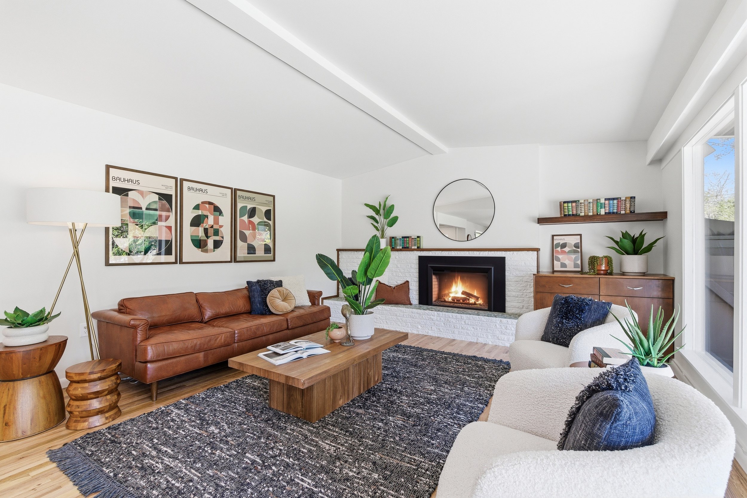 Bright living room with white walls, a fireplace, brown leather sofa, white armchair, wooden coffee table, green plants, artwork on walls, and large window letting in natural light.