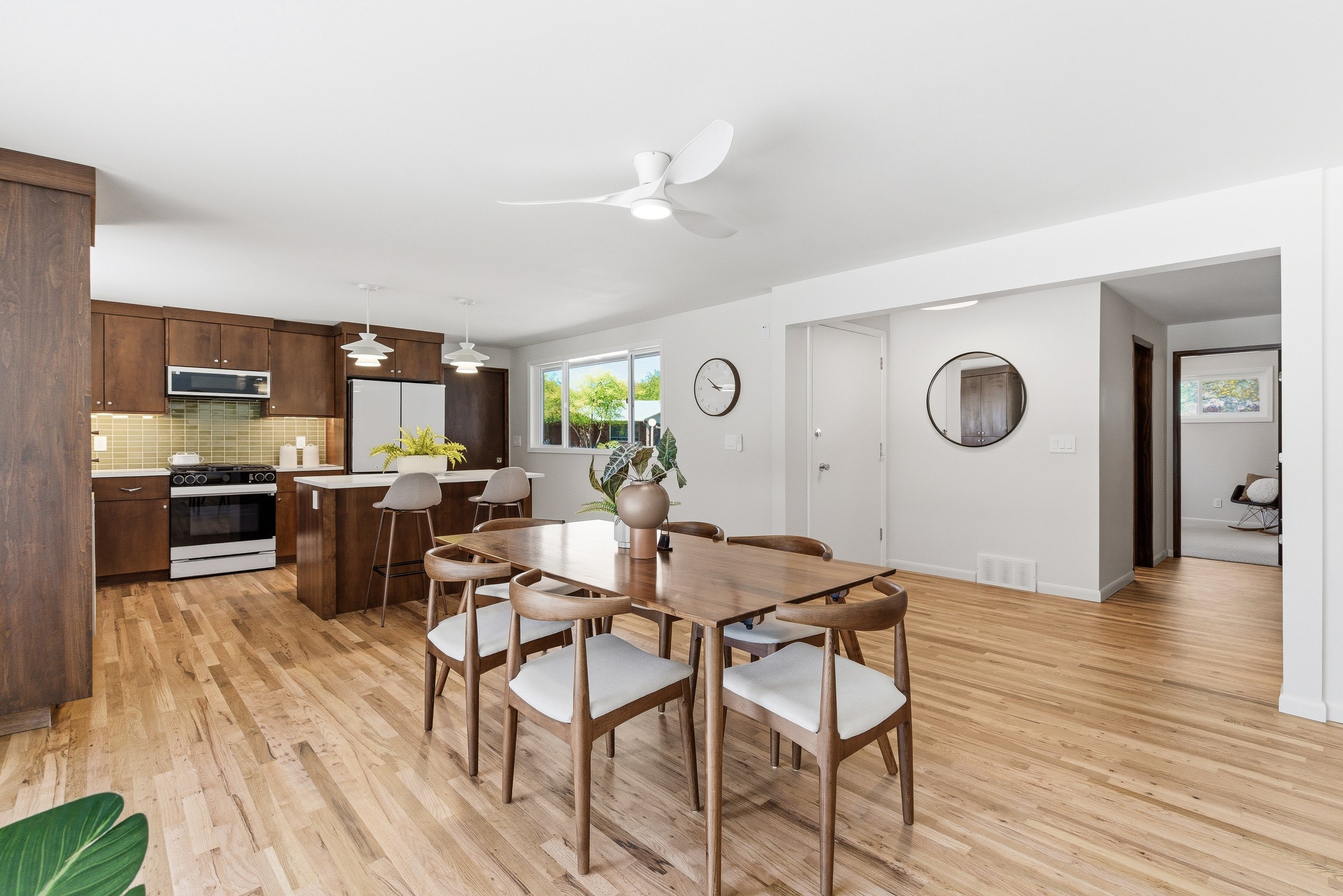 Open-concept dining and kitchen area with wooden flooring, white walls, modern lighting, a round dining table with six chairs, a kitchen with dark wood cabinets, a white refrigerator, and green backsplash tiles, sunlit through large windows. A ceilin