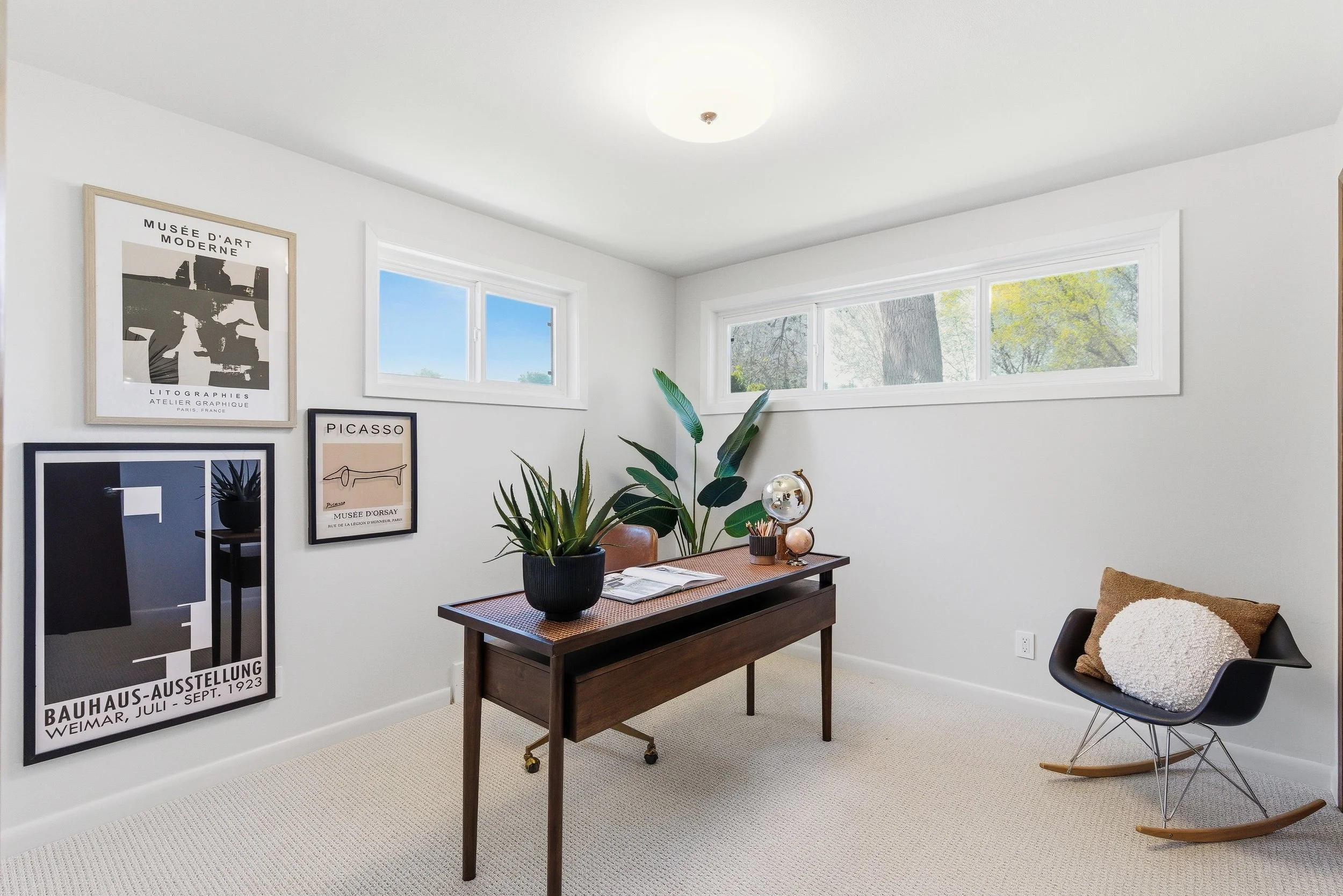A modern home office with white walls and a beige carpet. The wall features a gallery of framed art and posters, including one titled "Musée d'Art Moderne" and another for "Picasso." A dark wooden desk with a plant, open book, and decorative items is
