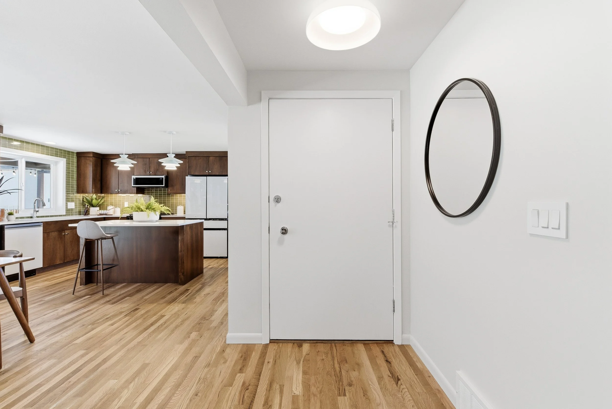 Modern kitchen and dining area with wooden floors, white walls, a white door, black oval mirror, and light fixtures.