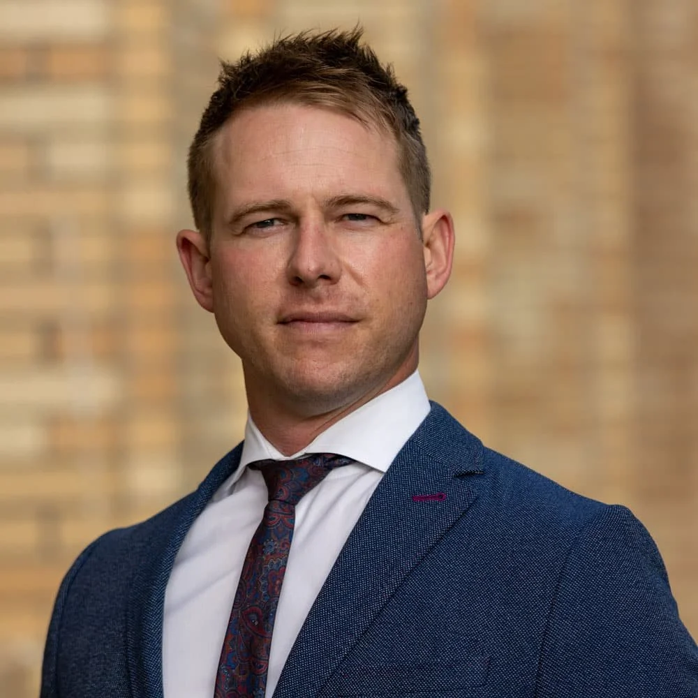A man in a navy blue suit and patterned tie standing outdoors with a blurred brick wall background.