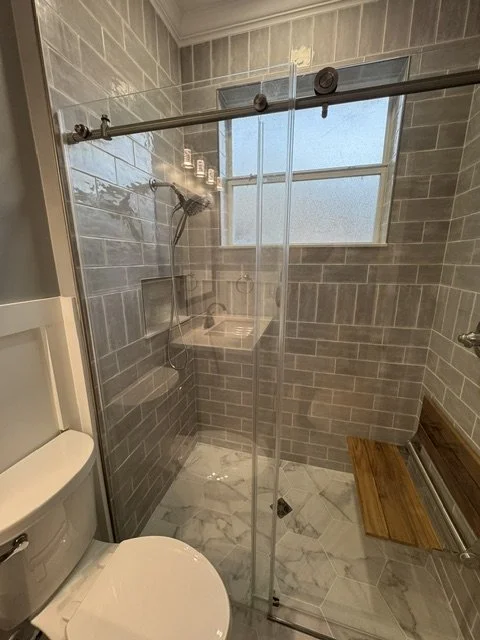 Bathroom shower with glass door and gray subway tile walls, a small window, wooden bench, and a toilet to the left.