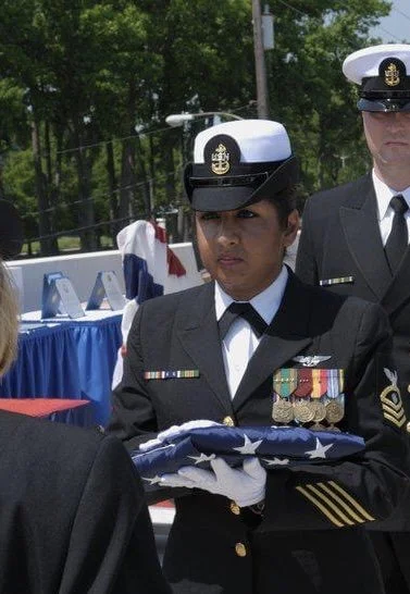 A female military officer in uniform holding a folded American flag during a formal ceremony, with other uniformed personnel and tables in the background.