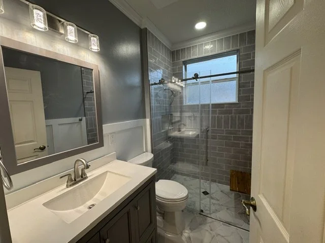 Bathroom featuring a white vanity with sink, mirror with lights, toilet, and a glass-enclosed shower with gray brick walls and a window.