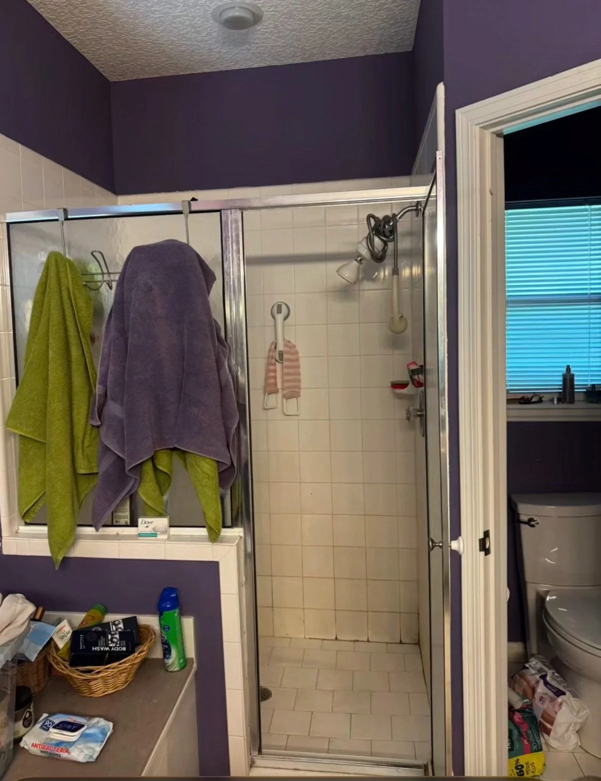 A bathroom with purple walls, a walk-in shower with two towels hanging on the door, and a window with closed blinds. The bathroom appears cluttered with toiletries and items on a counter and shelf.