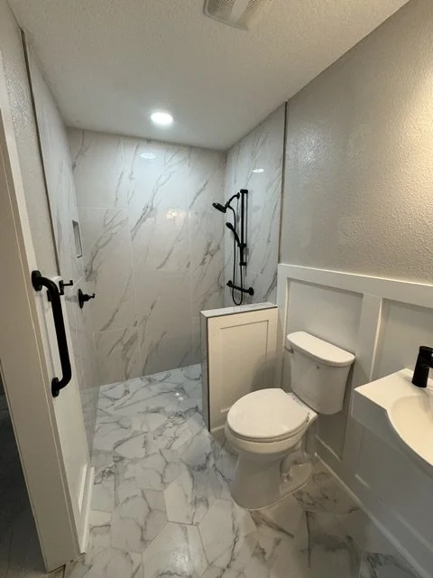 Small bathroom with a walk-in shower that has a black showerhead and black handrails, gray walls, and a white toilet with a small sink nearby.