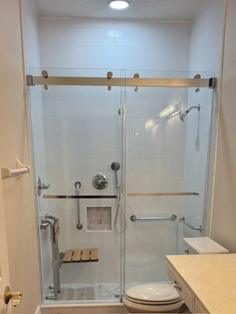 Bathroom with a walk-in shower having glass doors, a shower head, handrails, and a small wooden bench, along with a toilet and a sink.