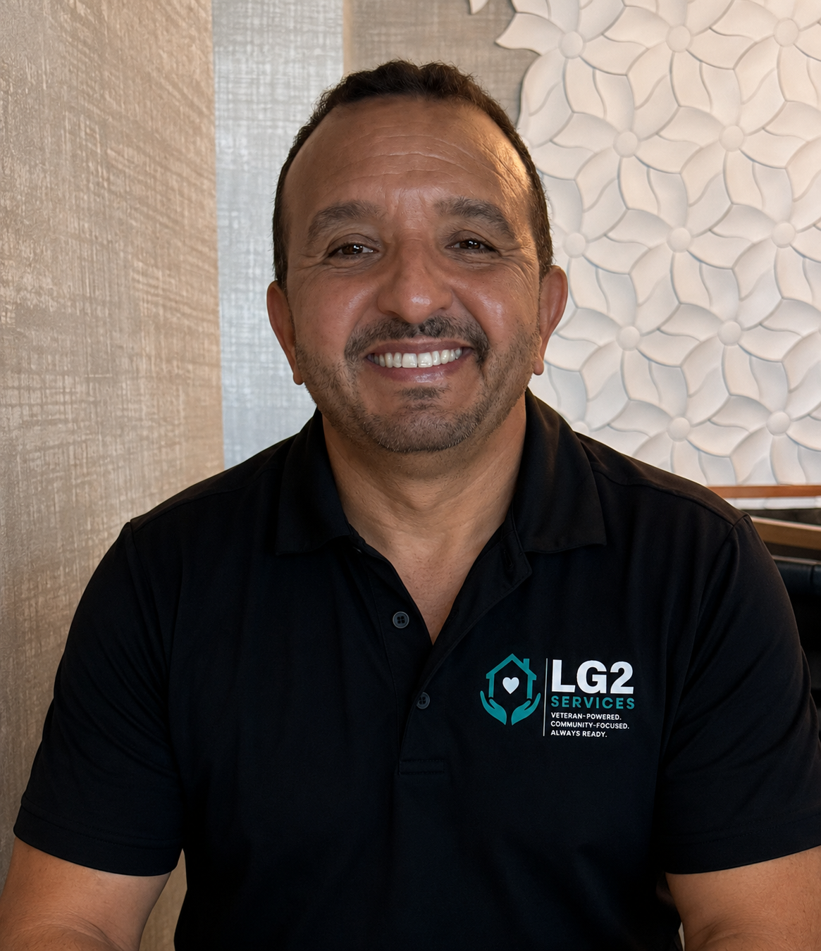 A smiling man with short dark hair and a beard, wearing a black polo shirt with the LG2 Services logo, sitting indoors against a textured wall and decorative white wall art.