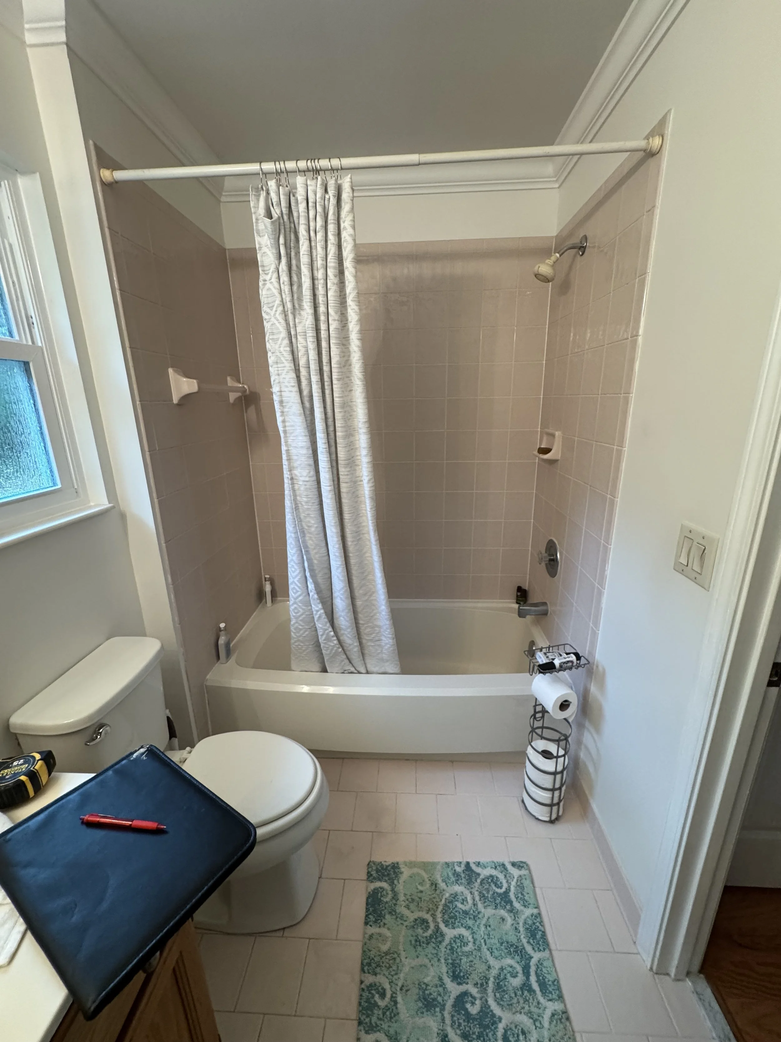 Bathroom with a bathtub and shower, beige tiles, a white curtain, toilet paper holder, and a window with natural light.