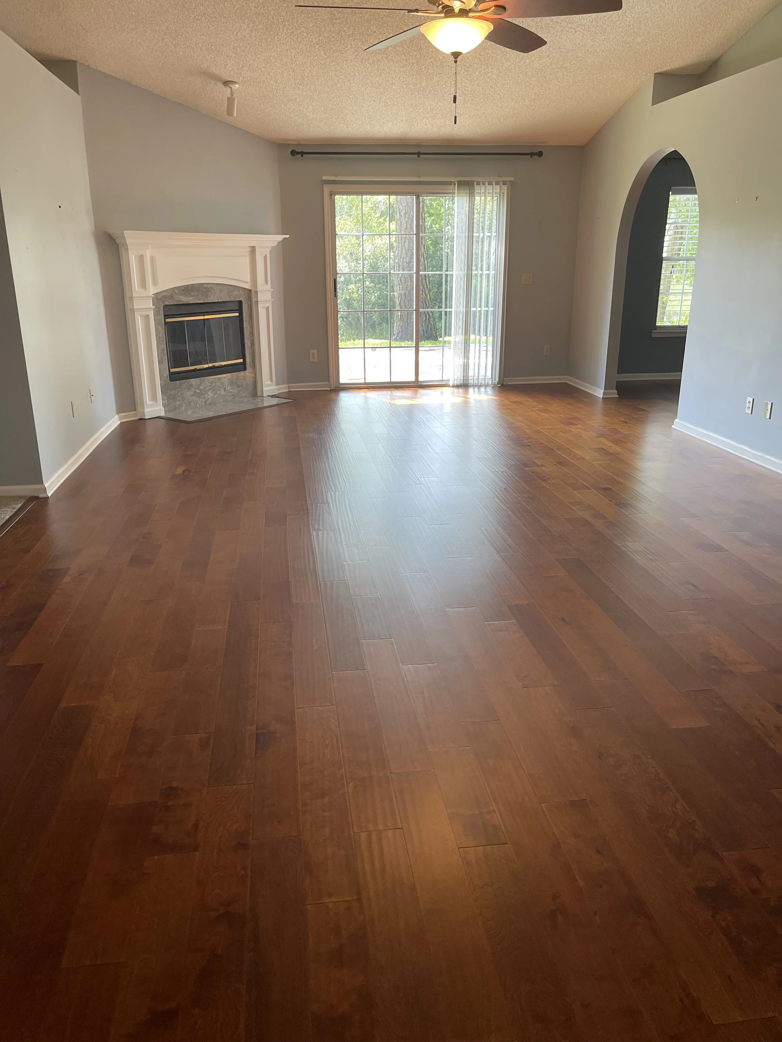 Empty living room with hardwood floors, a fireplace with white mantel, a sliding glass door leading outside, and an arched doorway. Ceiling fan with light fixture.