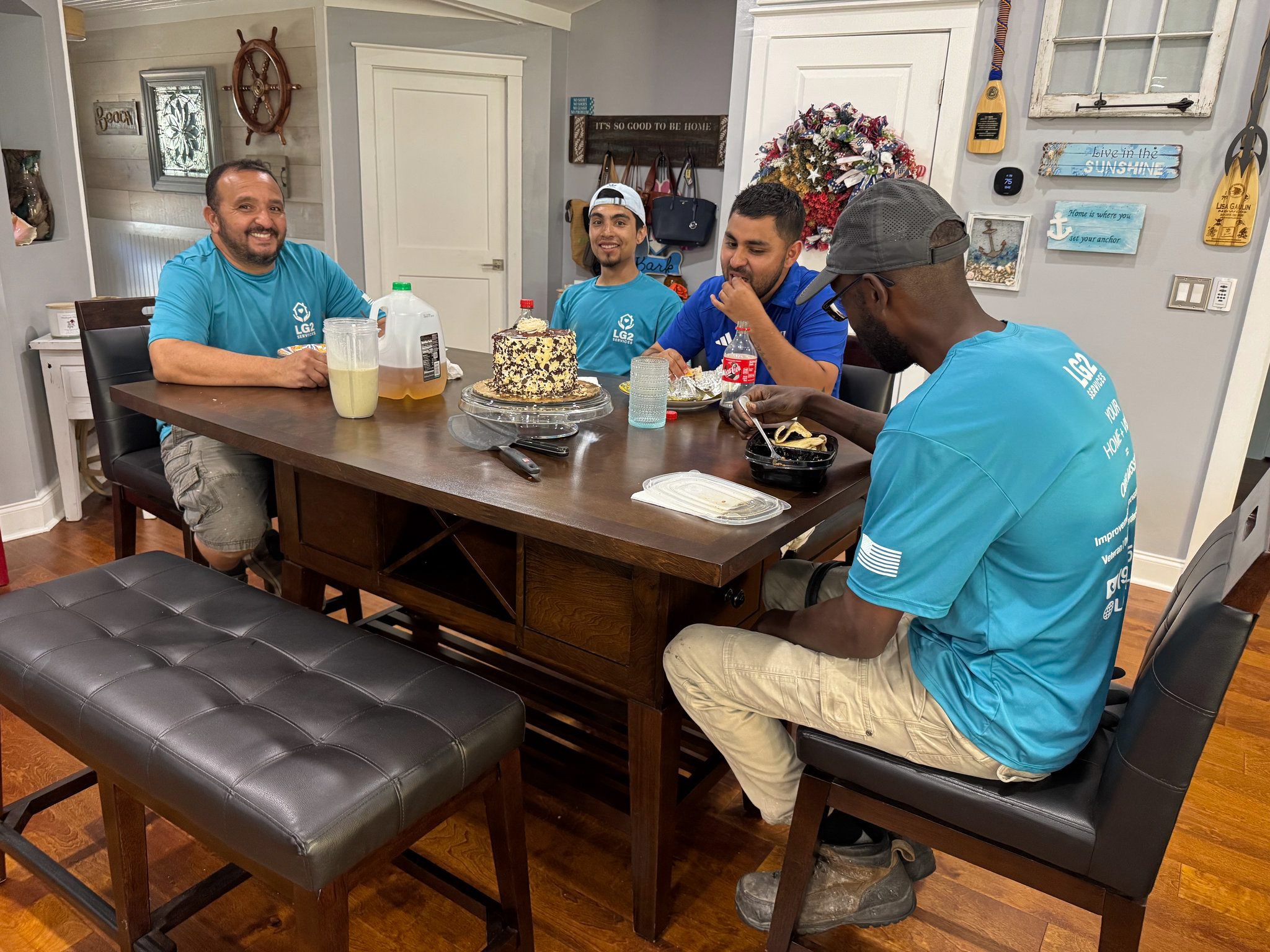 Four men sitting around a dining table with a cake, drinks, and food in a decorated home interior, celebrating.