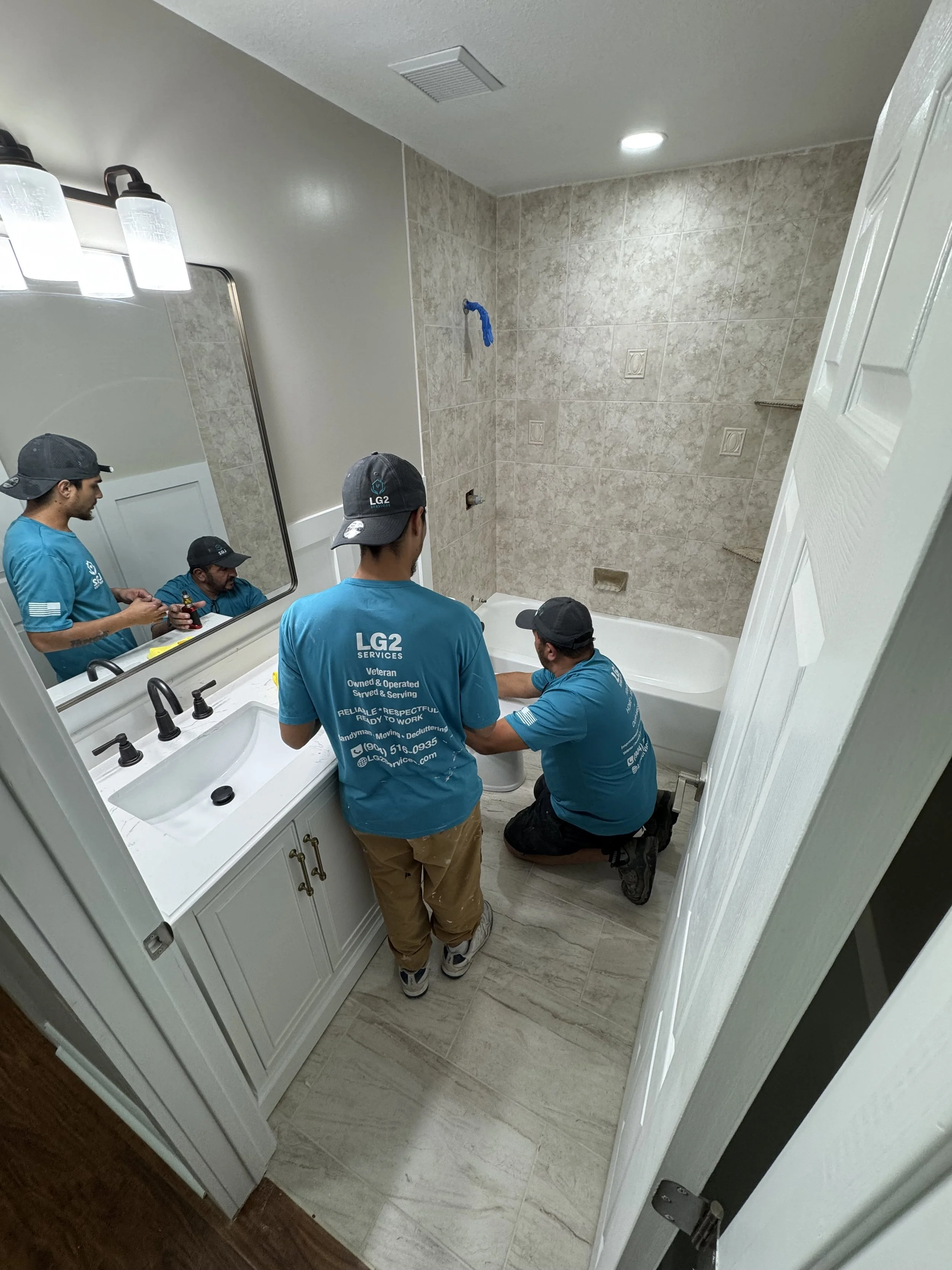 Four workers are installing or repairing a bathroom, with two of them standing and two kneeling, working on the bathtub area. One worker is sitting on the edge of the bathtub. The bathroom has a mirror, a vanity with two sinks, and wall tiles.
