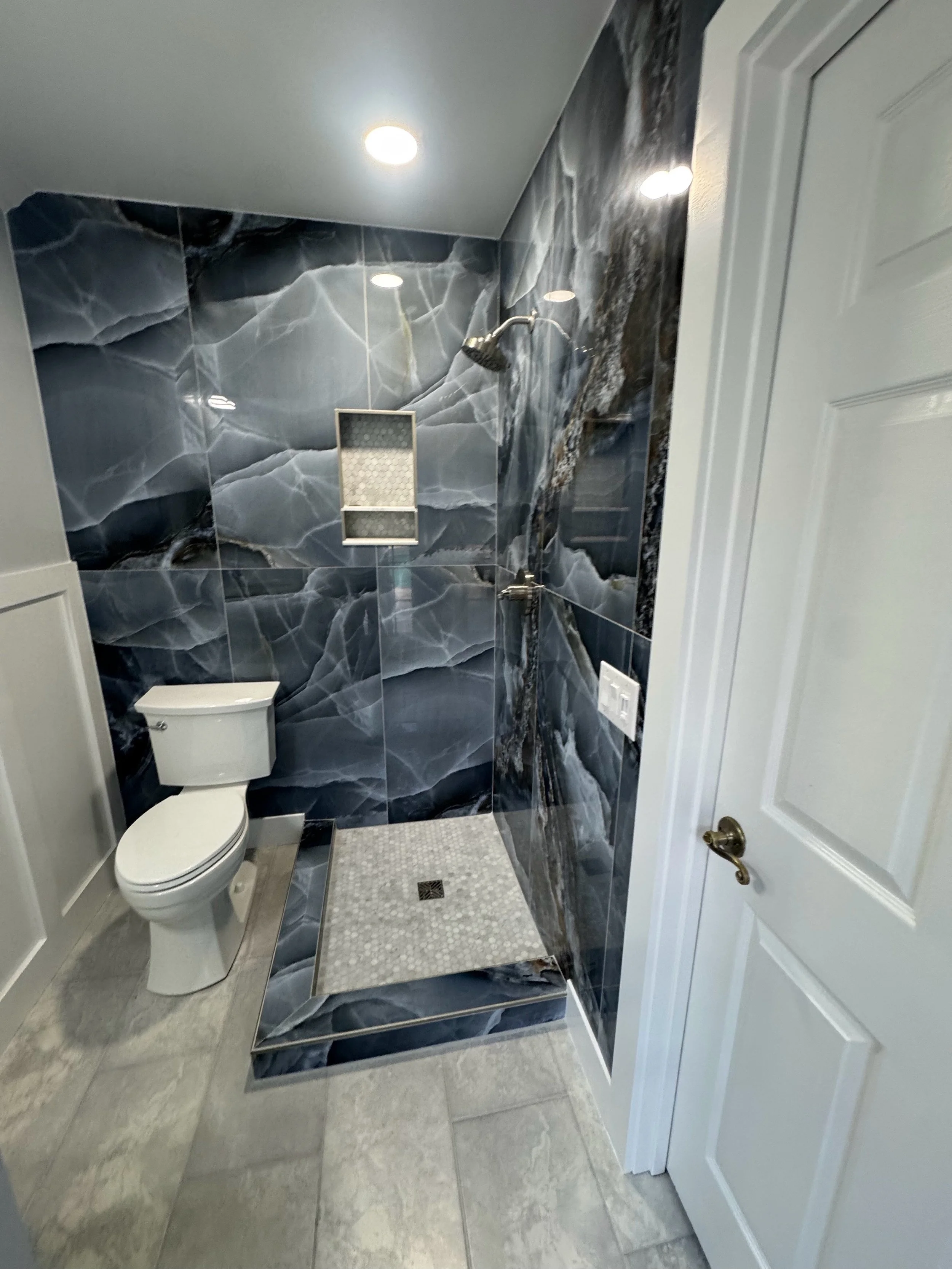 A modern bathroom with blue marble walls, a toilet, a standing shower with a rainfall shower head, a small tiled shower floor, and a ceiling-mounted light fixture.