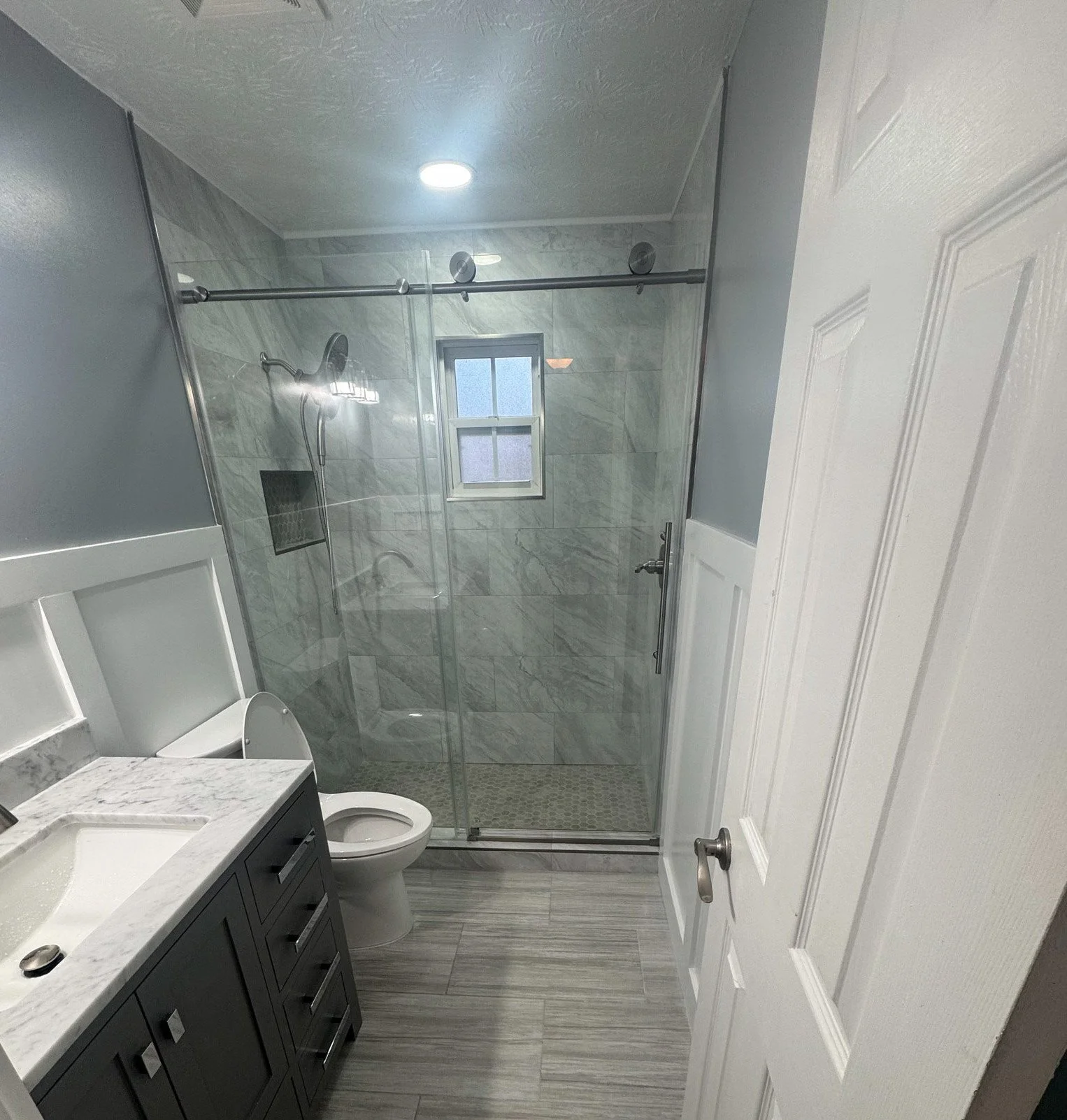 Modern bathroom with marble tile shower, glass door, small window, vanity with dark cabinet and white countertop, toilet, gray flooring, and white door.