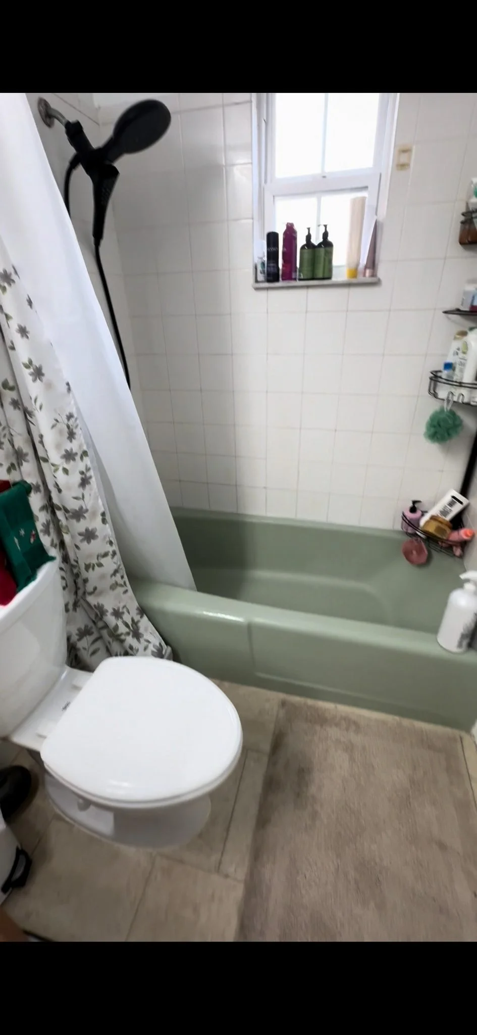 A small bathroom featuring a toilet, a bathtub with a showerhead, a window with several bottles on the sill, and various toiletries and cleaning supplies around the bathtub area.