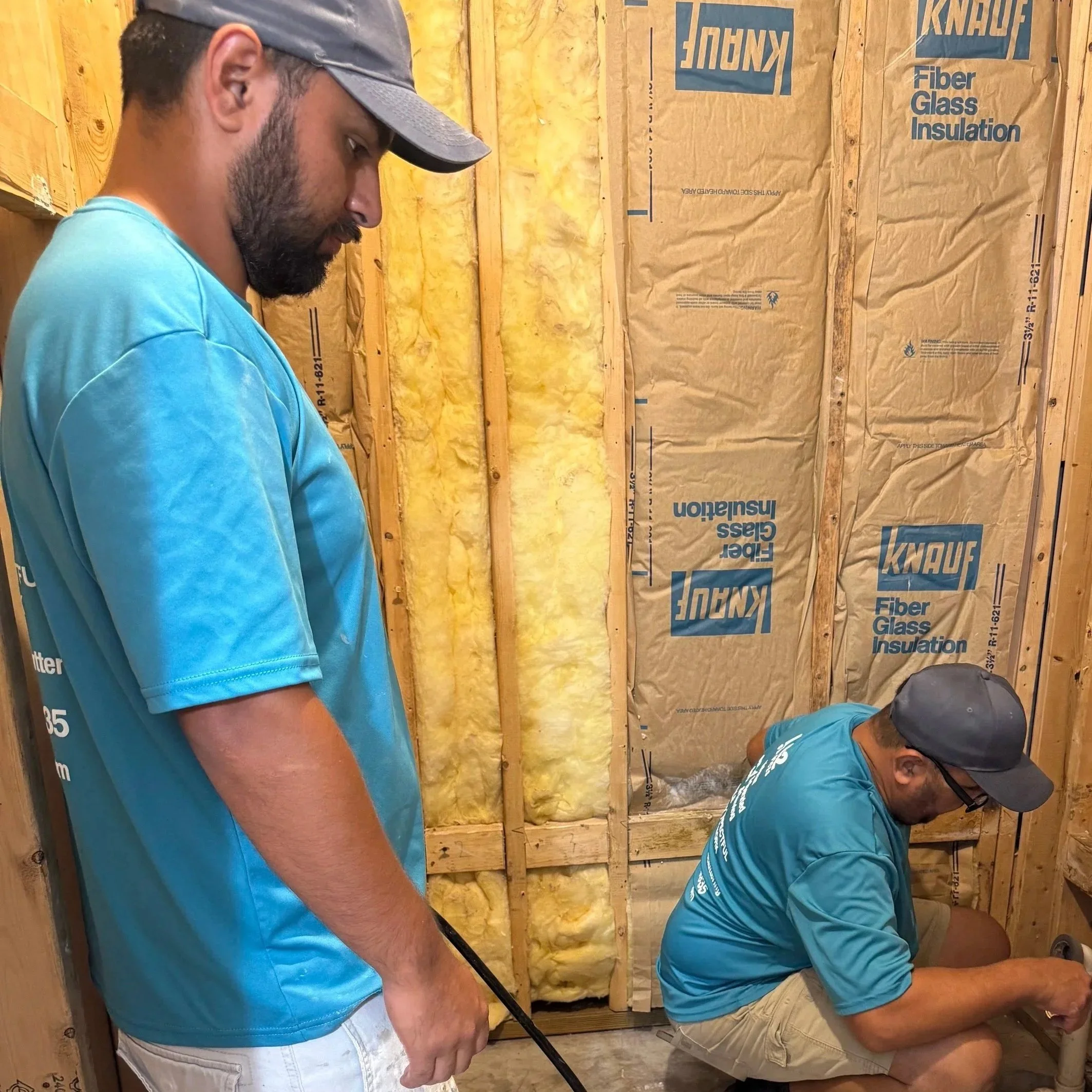 Two men wearing blue shirts working inside a structure with wooden framing and insulation in the walls.