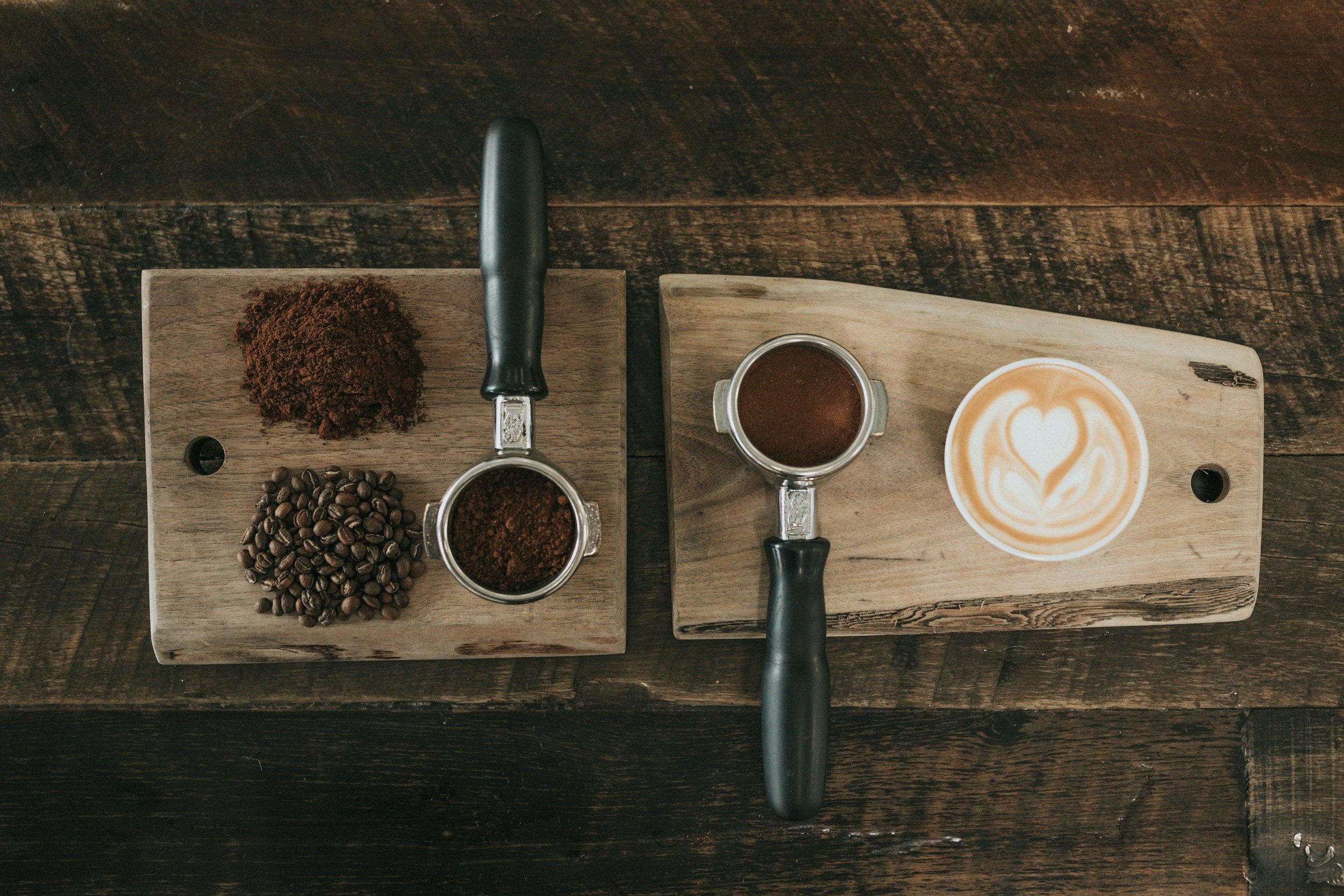 Top-down view of two wooden boards, one with ground coffee, coffee beans, and a coffee tamper, and the other with a cup of brewed coffee and a latte with foam art, on a dark wooden table.