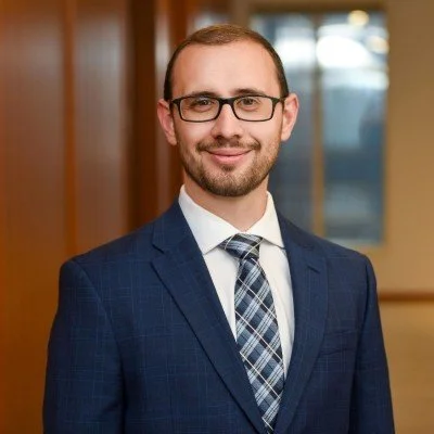 A man with glasses, short hair, and a beard wearing a navy blue checked suit, white shirt, and plaid tie, standing indoors in front of a wooden wall and window.