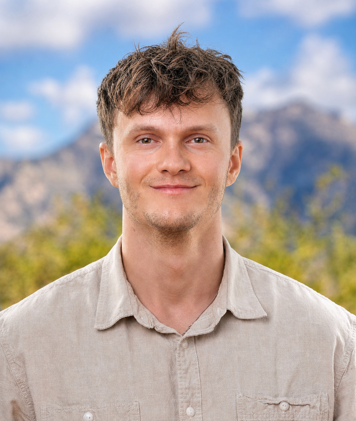A young man with light brown hair and light facial hair smiling outdoors with mountains and trees in the background.