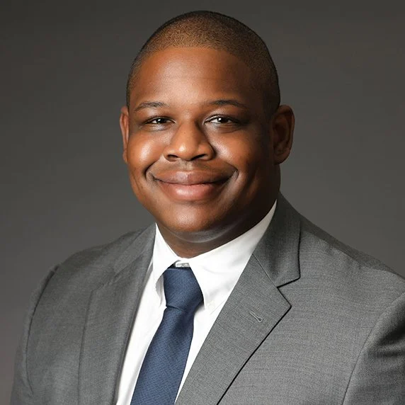 Professional portrait of a man in a gray suit, white shirt, and navy tie, smiling at the camera against a gray background.