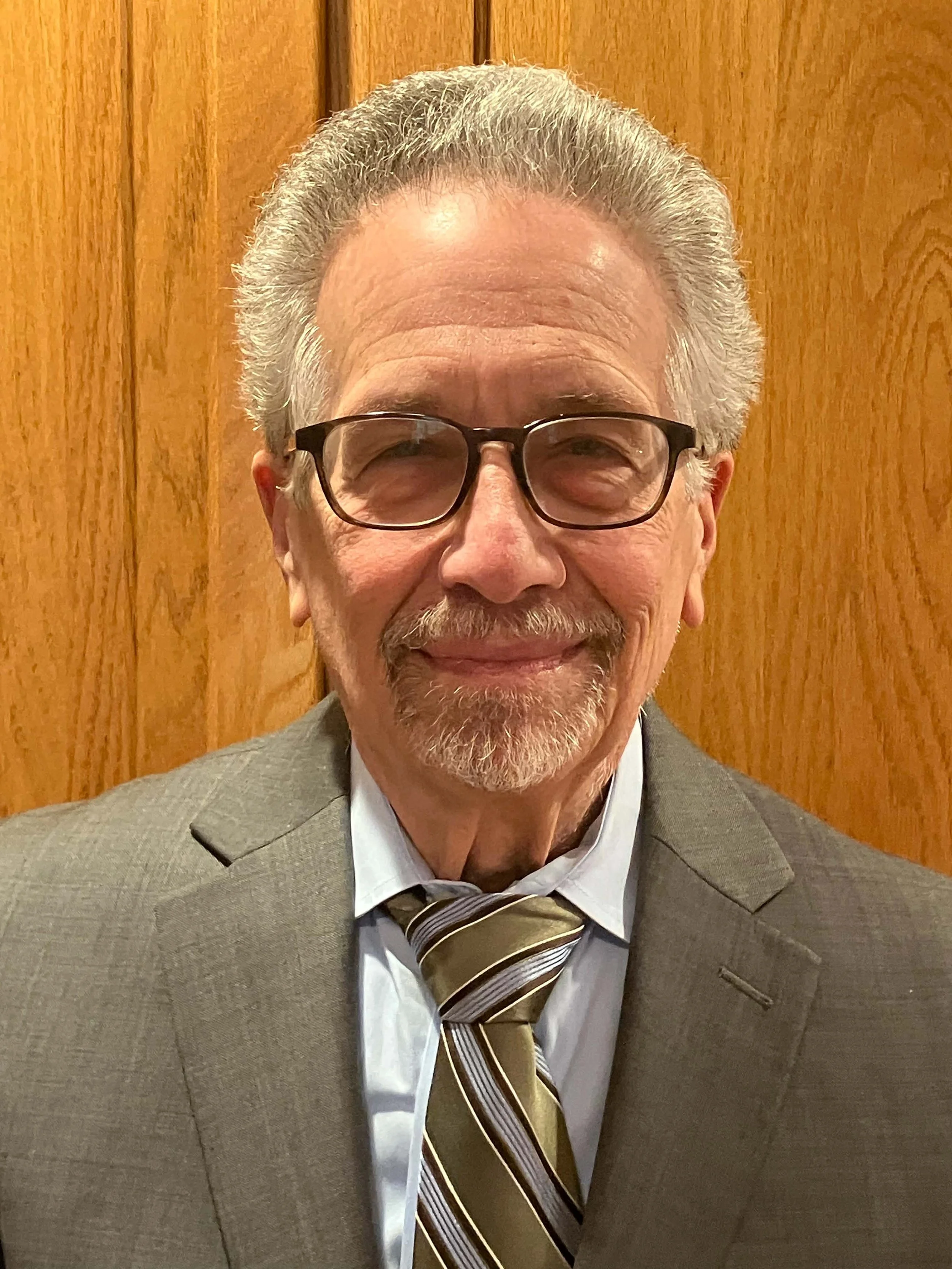 A smiling older man with gray hair and beard, wearing glasses, a gray suit, white shirt, and striped tie, standing in front of a wooden background.