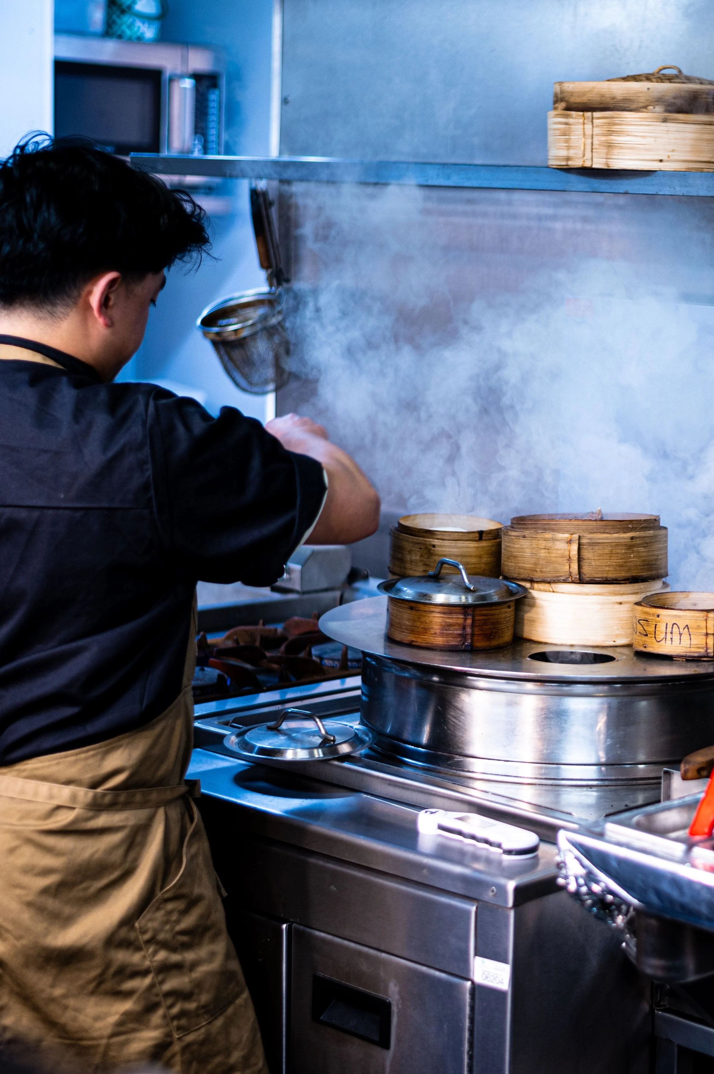 A person wearing an apron is working in a kitchen with steam rising from a steaming basket on a stove, and several other steaming baskets placed nearby.