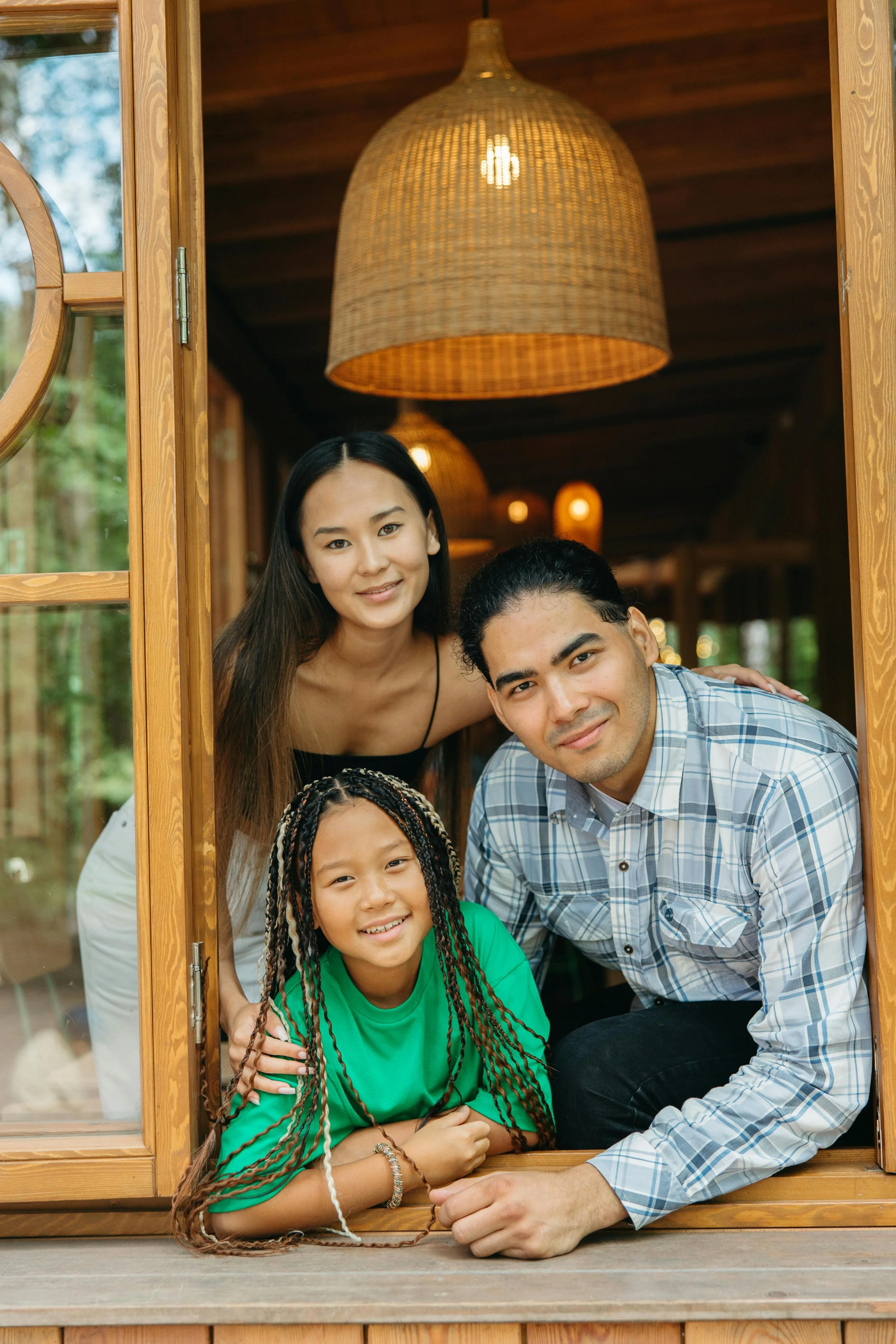 A diverse family of four peeking out of a wooden window, smiling, with green and wooded surroundings visible in the background.