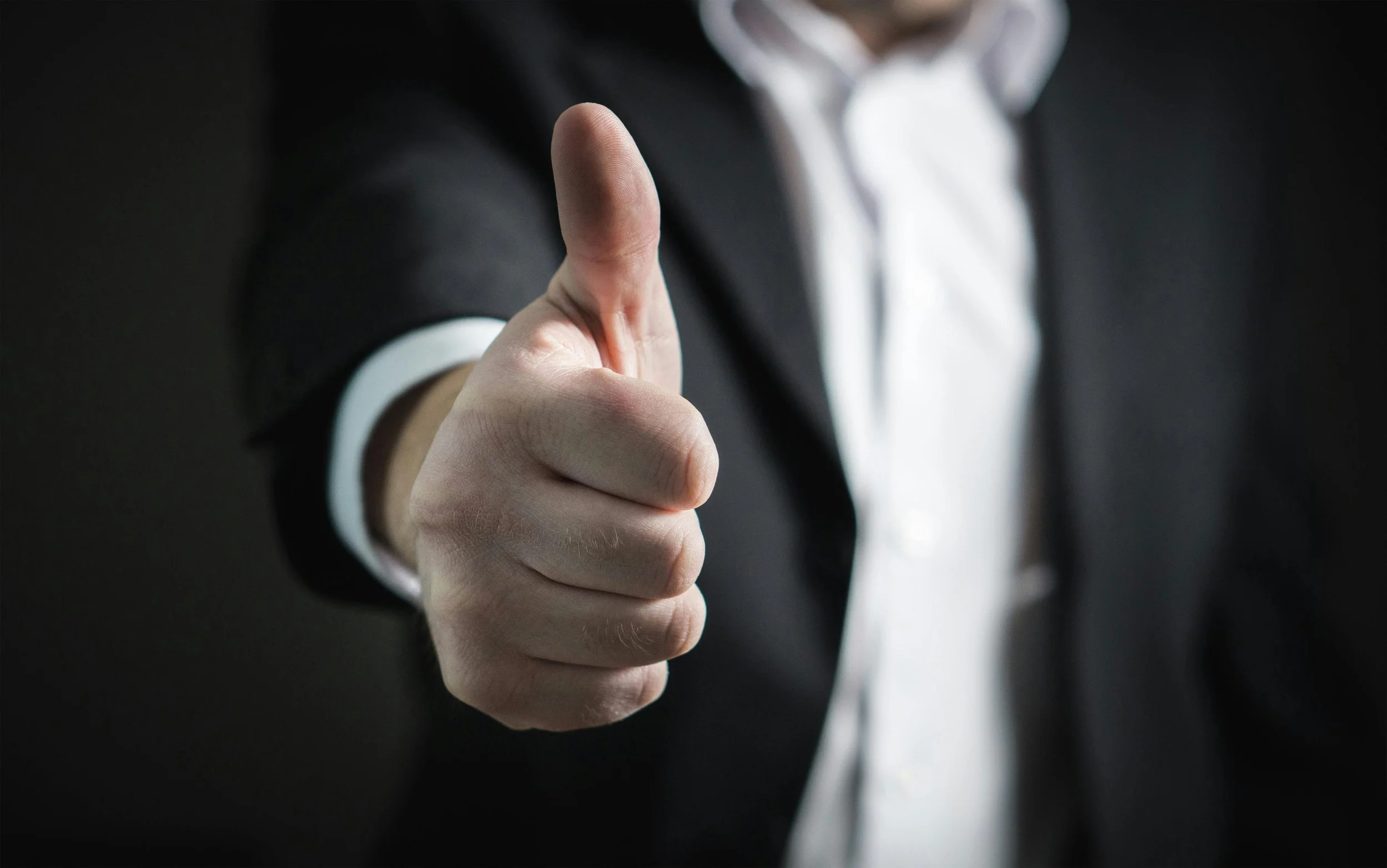 Person in a business suit pointing directly at the camera with a dark background.