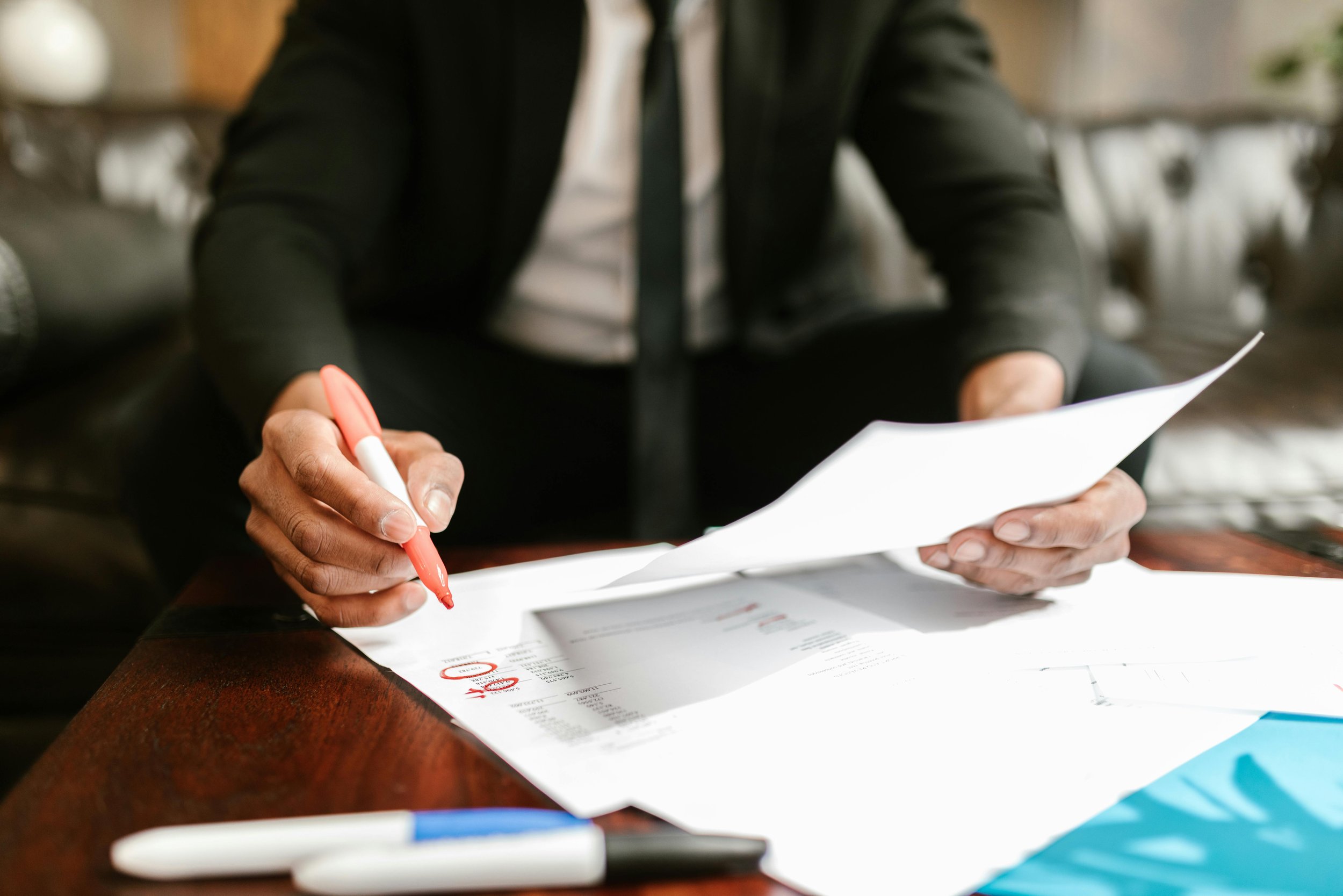 Person in a black jacket and tie sitting at a table, holding a red pen and filling out or reviewing a document, with a white and blue pen lying on the table, and a second sheet of paper with printed information and red markings.