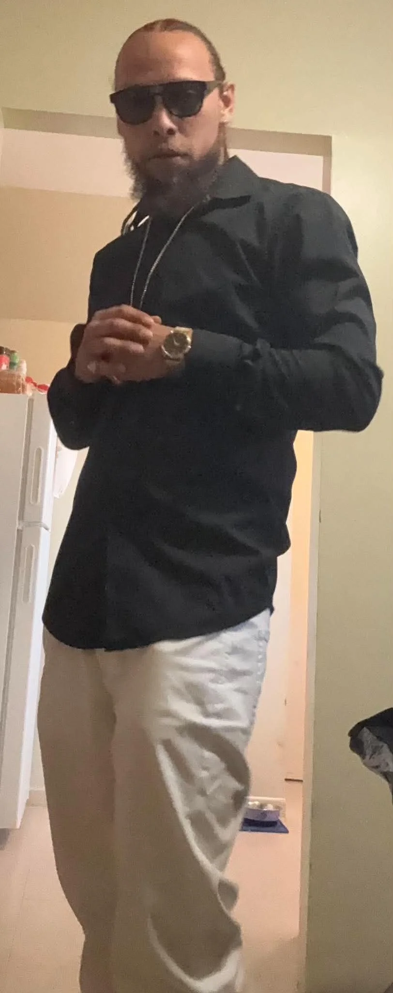 A man with sunglasses, a necklace, a watch, and a beard, wearing a black shirt and beige pants, standing in a room with a kitchen in the background.