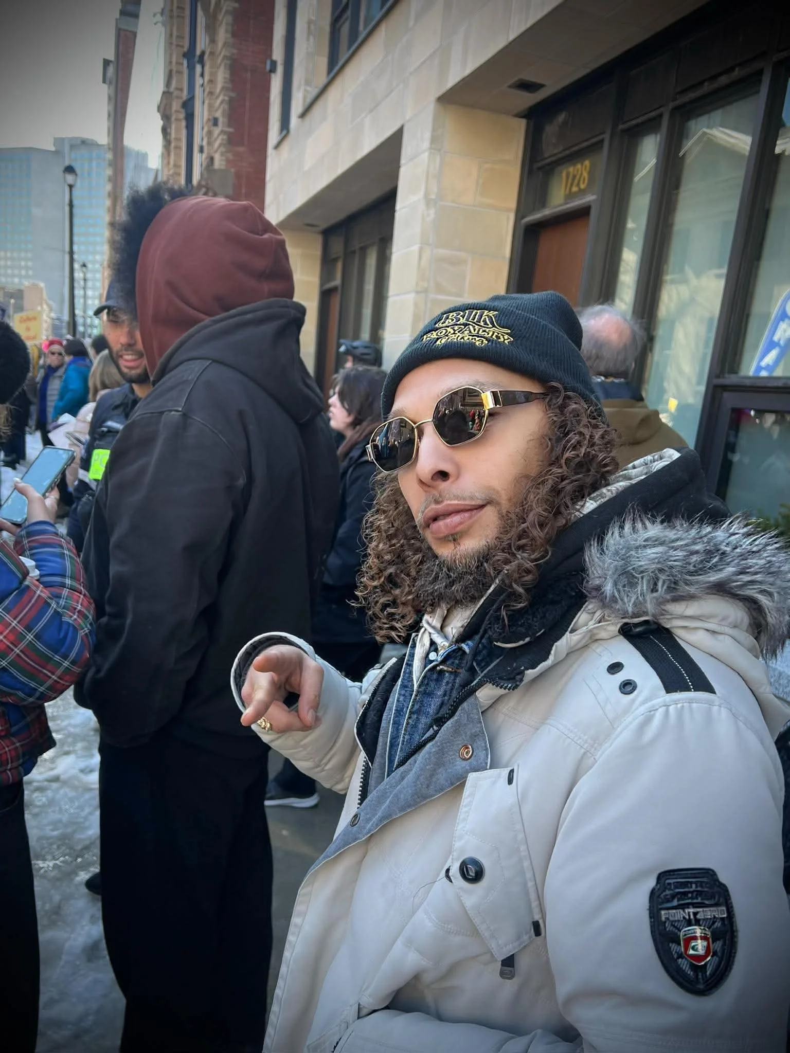 A man with curly hair and a beard wearing sunglasses, a black beanie, and a beige jacket with patches on the sleeve, standing in a crowd outside a building.