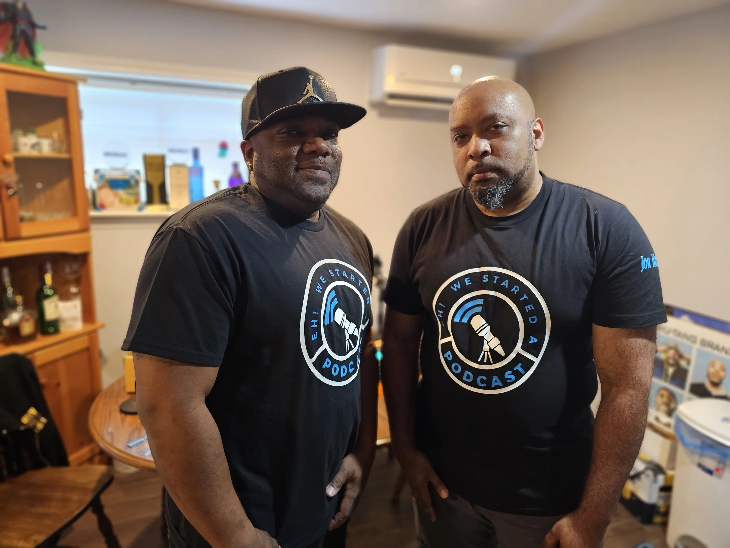 Two men wearing black T-shirts with a podcast logo stand indoors, looking at the camera. The man on the left wears a black cap with a Jordan logo, and the man on the right is bald with a beard. Background includes a wooden cabinet with bottles and a 