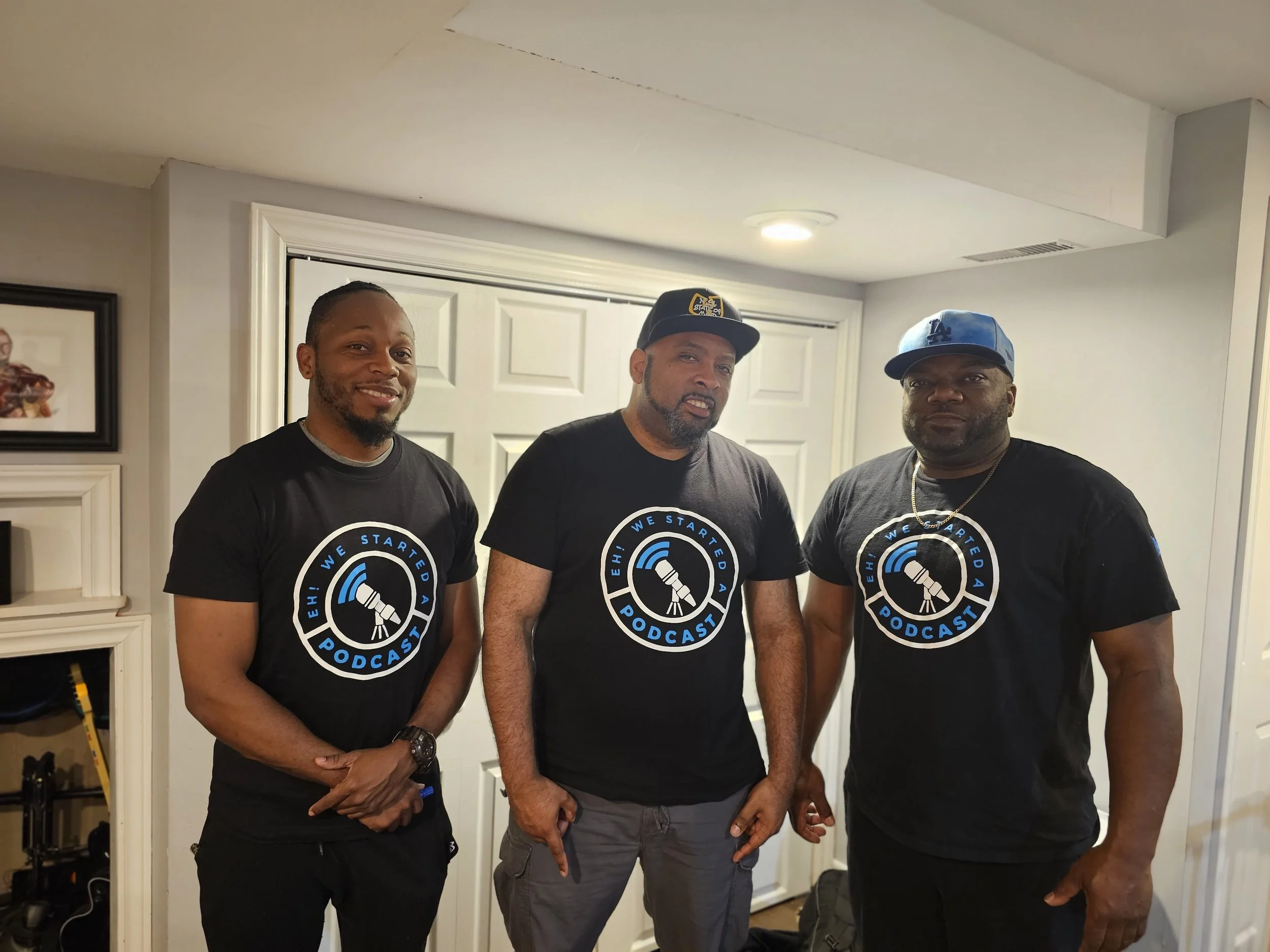 DJ Laton, Jon Kizzy and King Tygga wearing black T-shirts with a podcast logo, standing indoors in front of a white door, smiling and looking at the camera.