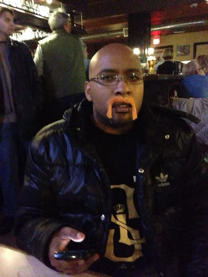 A man with glasses sits at a table in a dimly lit restaurant or bar, wearing a black leather jacket and a shirt with a large letter 'B' on it. He has a bald head, orange beard and mustache makeup, and is holding a phone in his right hand. In the back