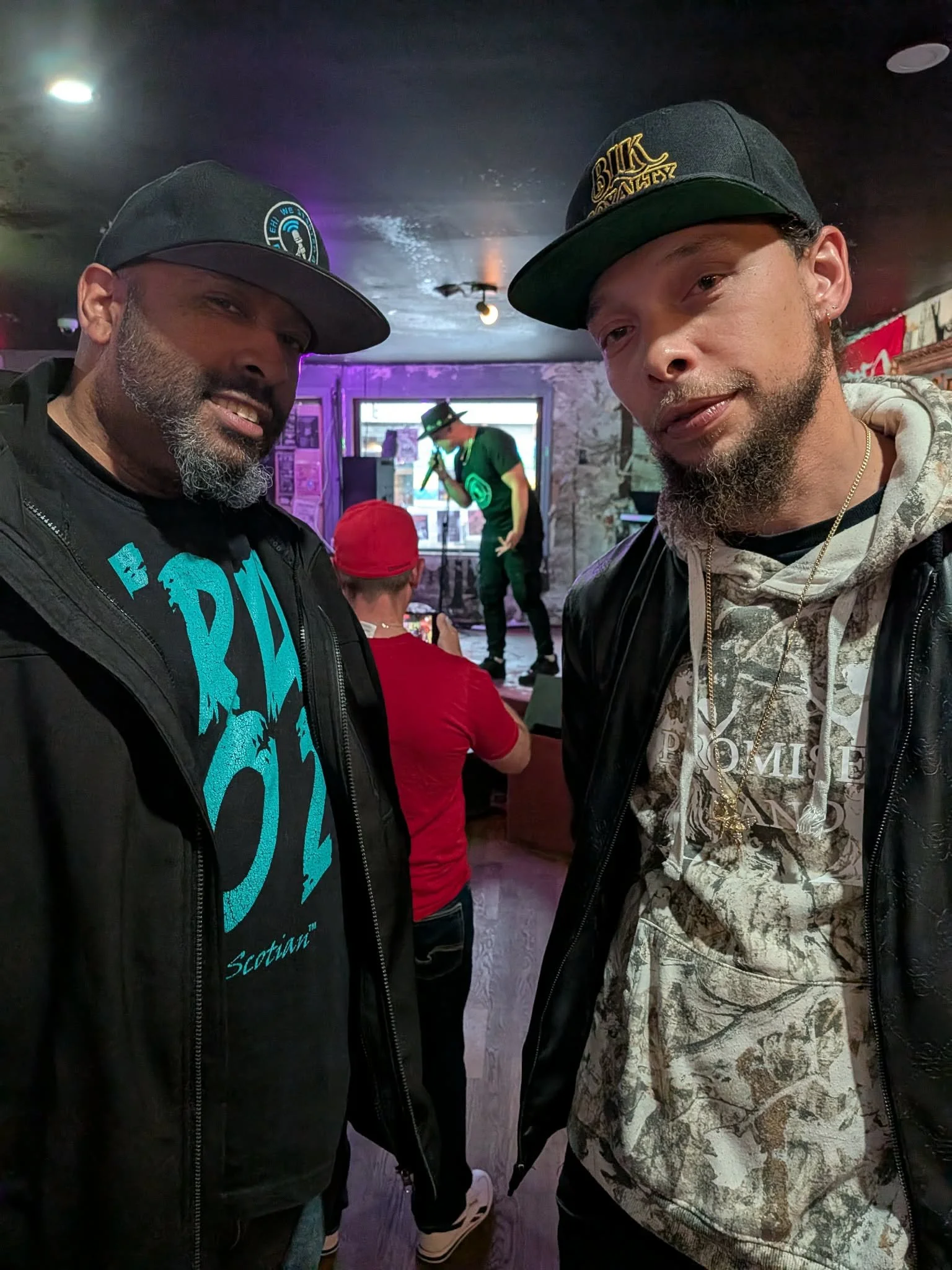 Jon Kizzy and Ray Rey wearing black caps and jackets taking a selfie in a dimly lit bar or club with Robbie G performing on stage in the background.