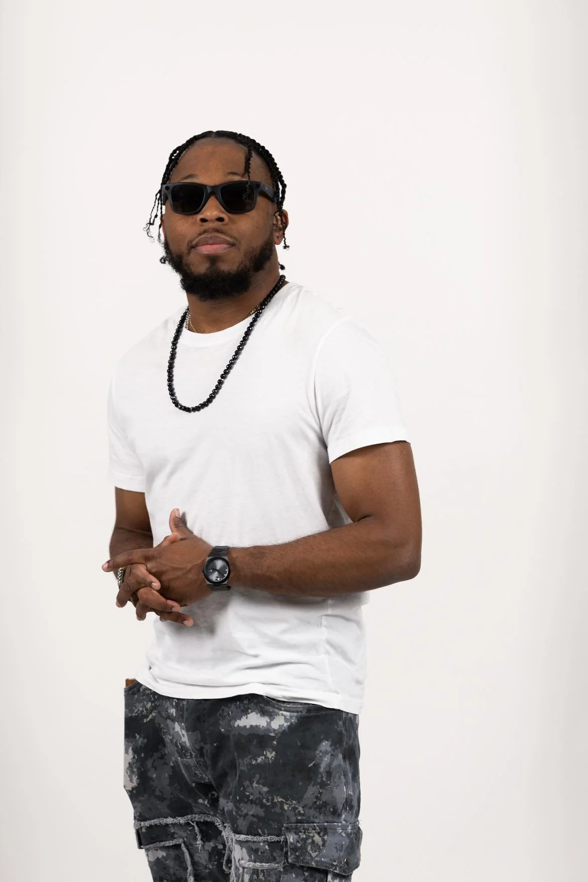 Man wearing black sunglasses, a white T-shirt, camouflage pants, a black wristwatch, a black beaded necklace, and a black bracelet, standing against a plain white background.