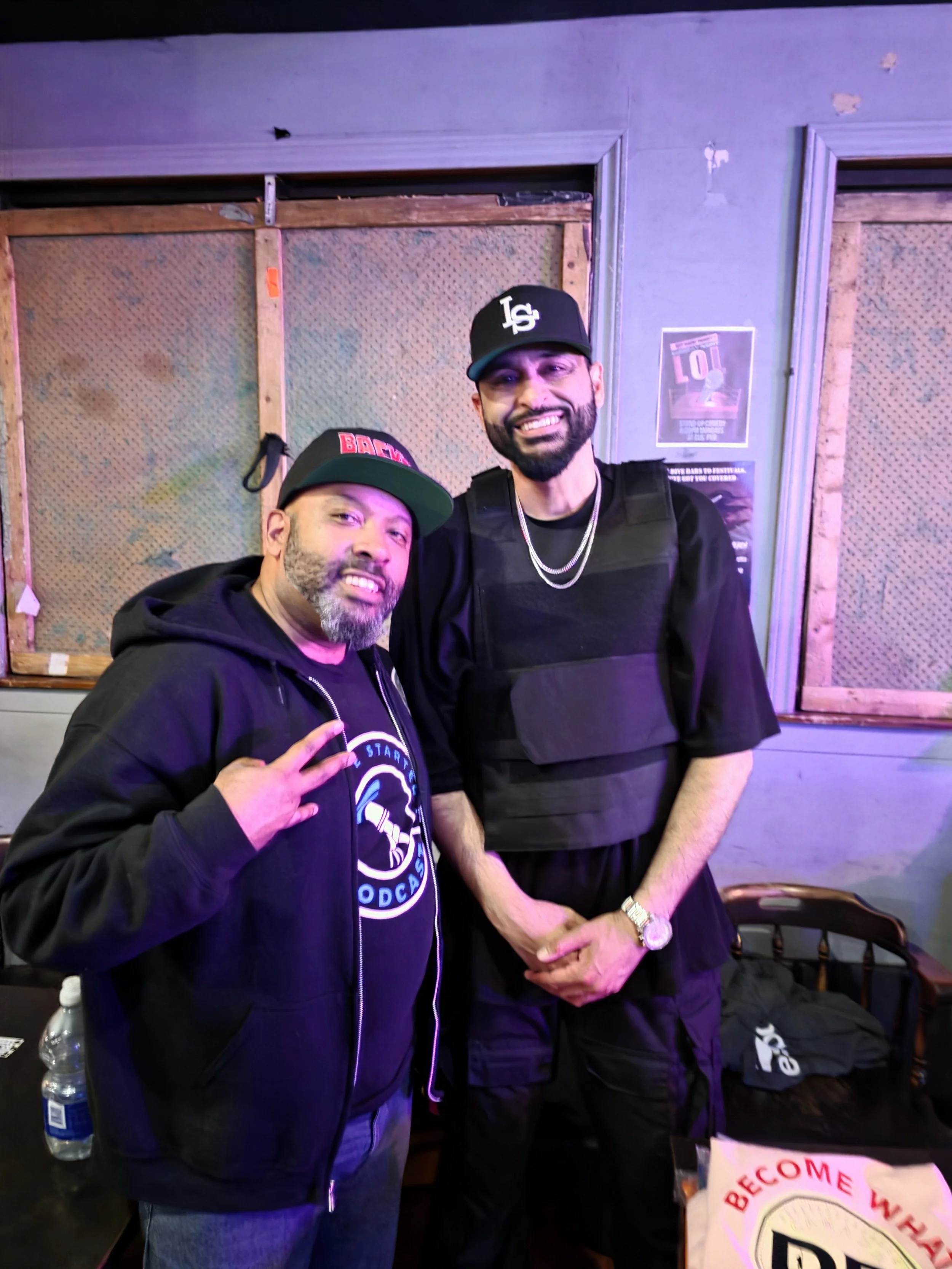 Two men standing together, smiling at the camera, in a room with purple lighting. The man on the left is wearing a black hoodie and a black cap with red and white lettering, making a peace sign with his right hand. The man on the right is taller, wit