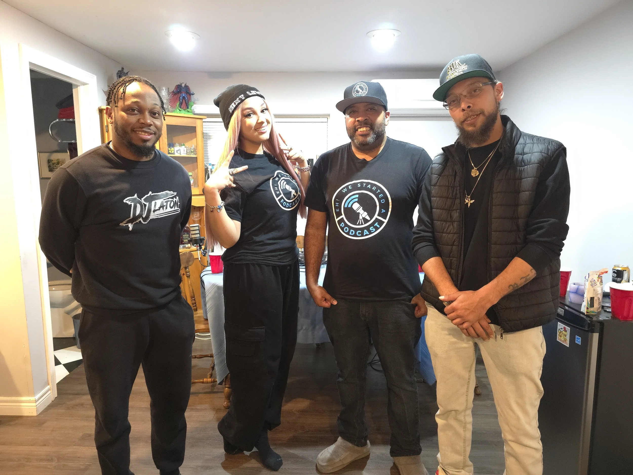 Four people standing indoors, posing for a photo. The person on the far left is smiling, wearing a black T-shirt. The second person is a woman with pink hair, wearing a black beanie and T-shirt, making a peace sign. The third person is a man with a b