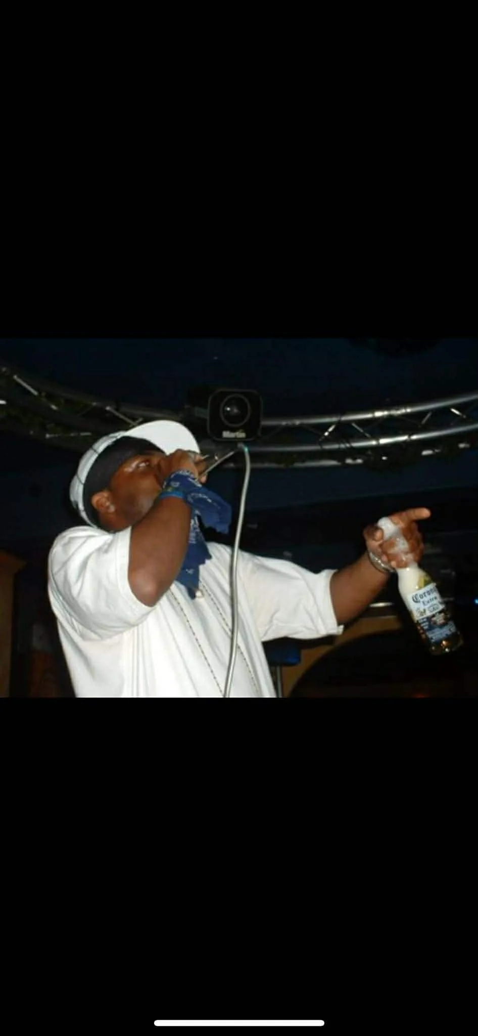 A man wearing a white shirt, white hat, and blue bandana sings or speaks into a microphone at a party or event. He holds a Corona beer bottle and is standing under a dark ceiling with business speakers above him.