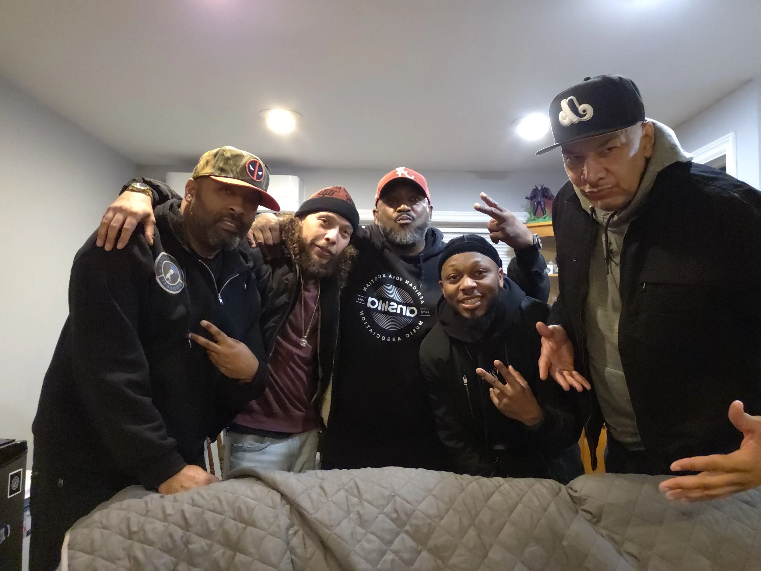 Jon Kizzy, Ray Rey, DJ Laton and Universal Soul posing around an object covered with a quilted blanket in an indoor setting.