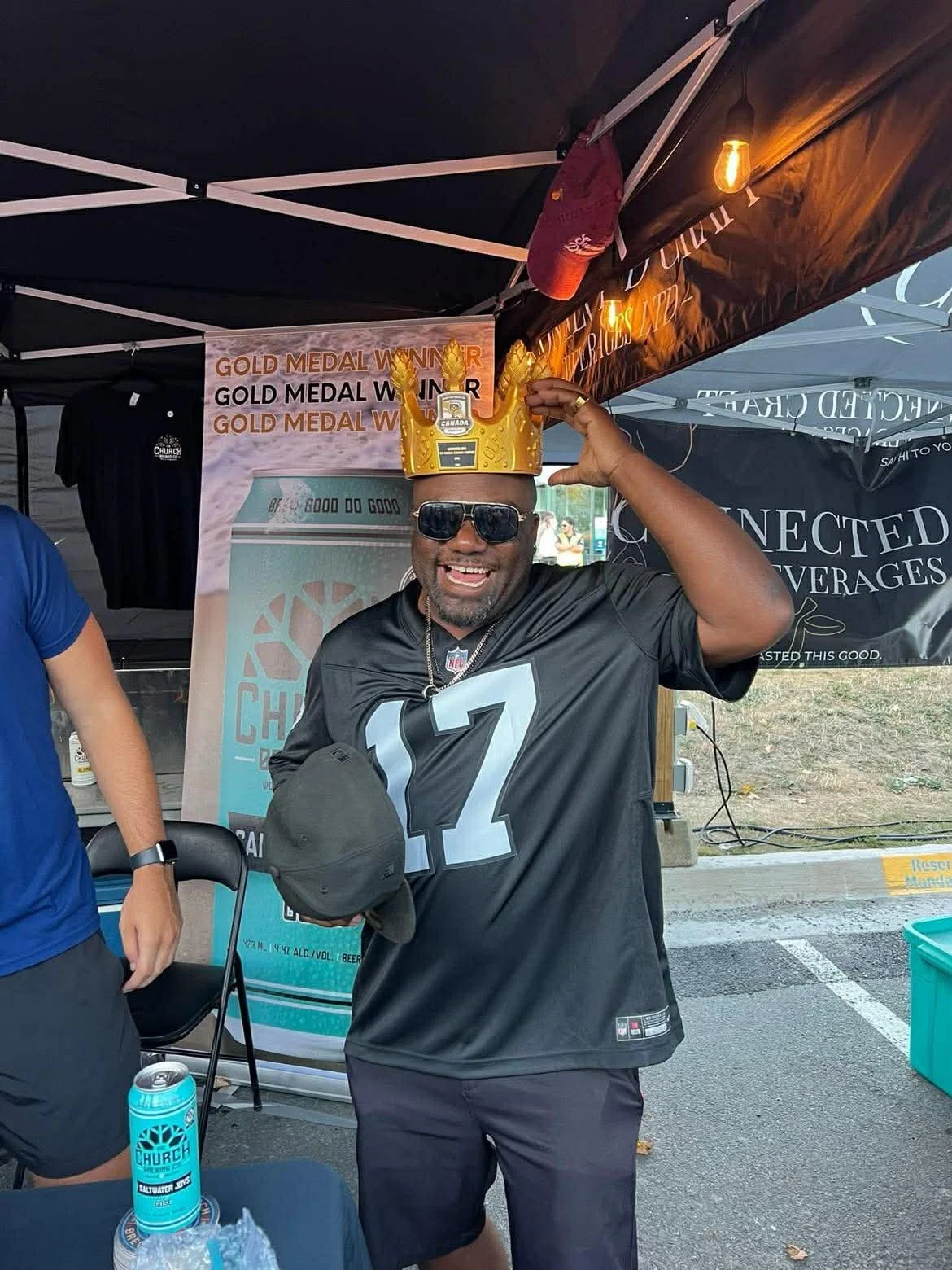 A man wearing a black sports jersey with the number 12, sunglasses, and a necklace is smiling while holding a black hat in one hand and adjusting a gold crown headpiece with the other hand, which is placed on his head. The background features tents a