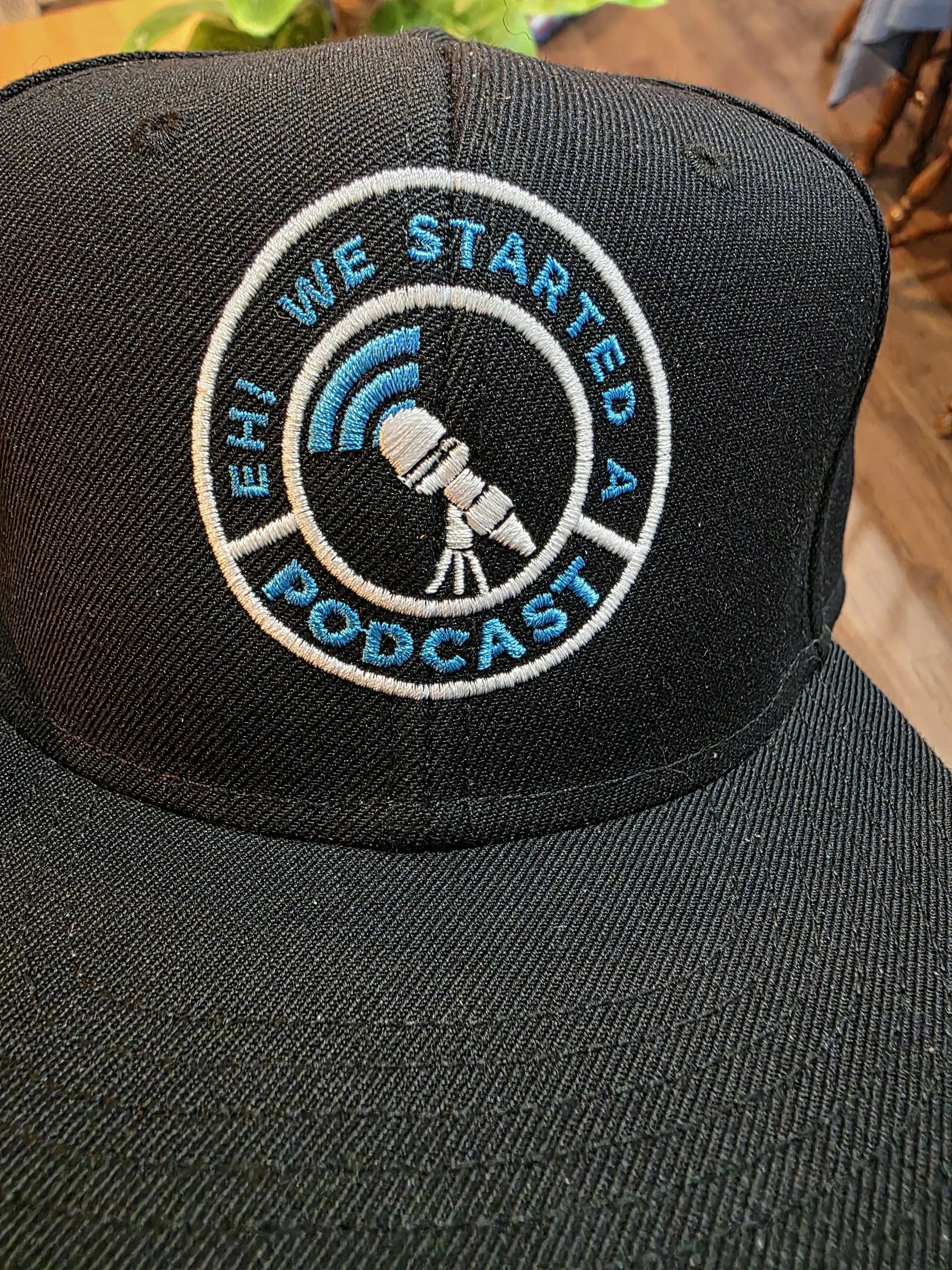A black baseball cap with an embroidered patch reading 'EH! We Started A Podcast' featuring a microphone and sound waves.