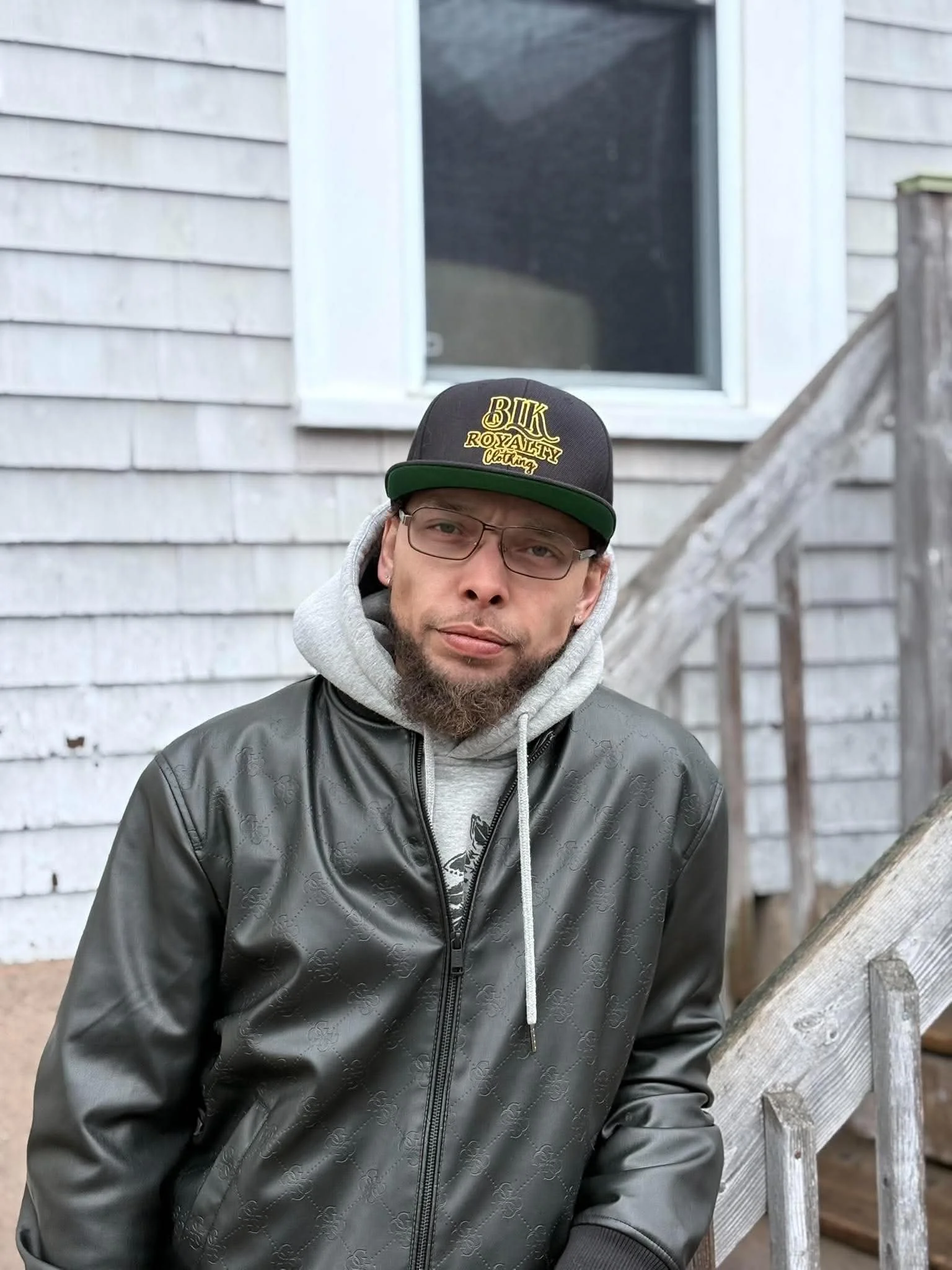Ray Rey wearing glasses, a black baseball cap with gold lettering, a gray hoodie, and a black jacket, standing outdoors in front of a house with gray wooden siding and a window.